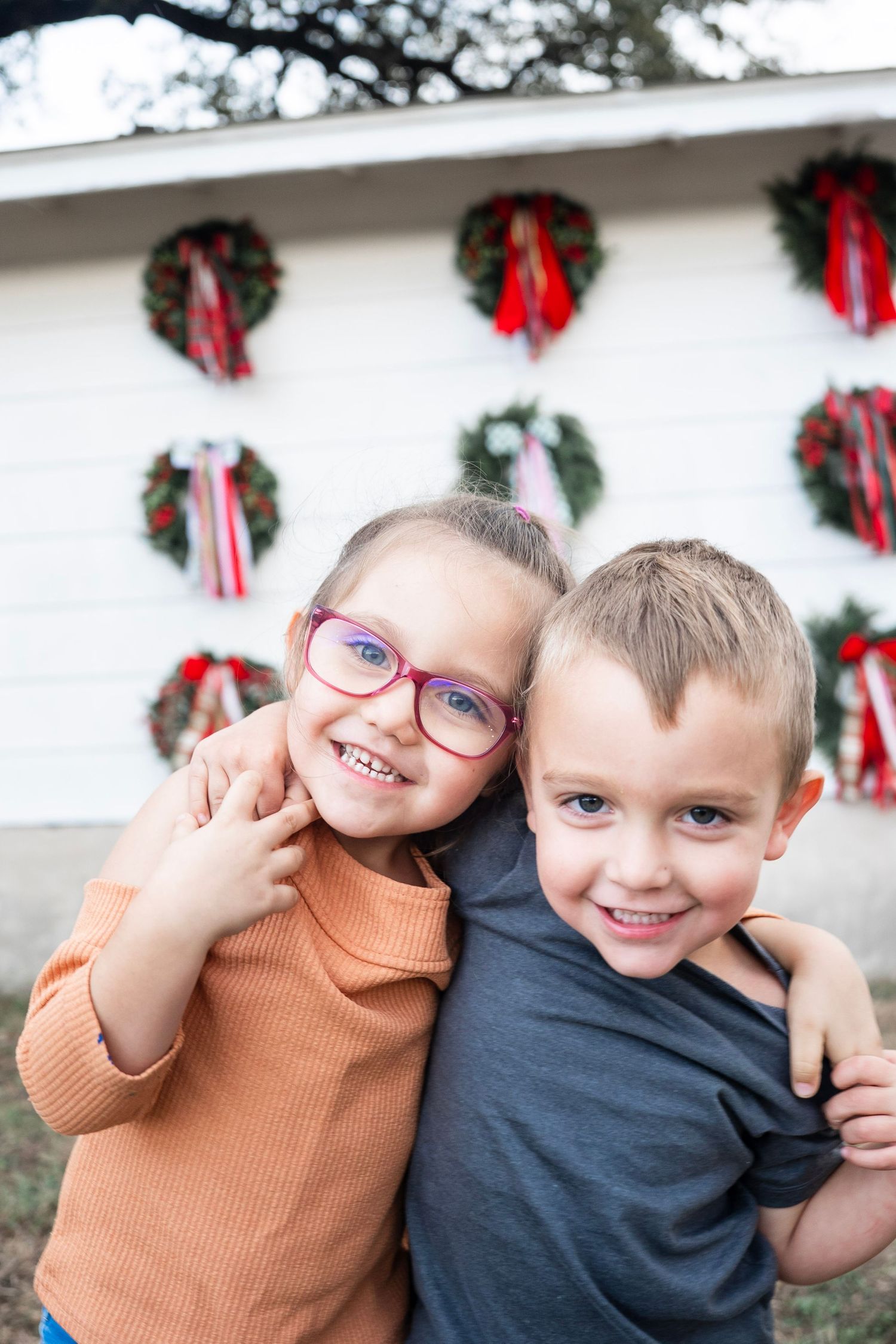 Two children in orange and navy sweaters pose playfully in front of a wall decorated with Christmas wreaths.