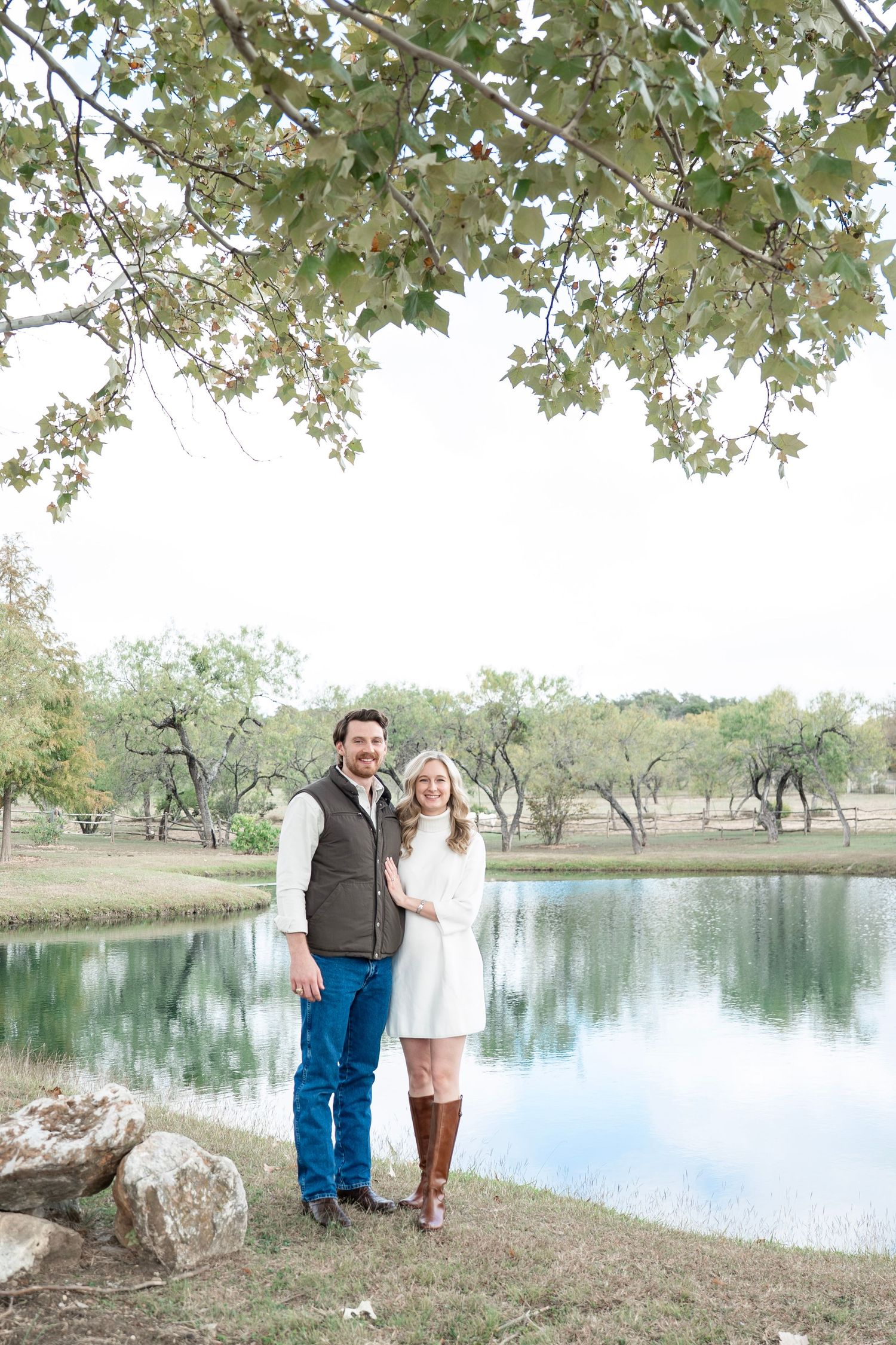 A couple stands together by a tranquil pond with overhanging tree branches and reflective water on a peaceful autumn day.