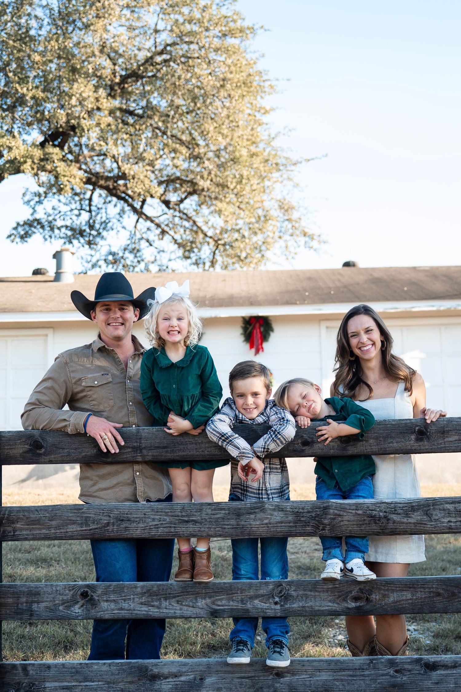 A family stands together at a rustic wooden fence during the holidays with a wreath visible on a building in the background.