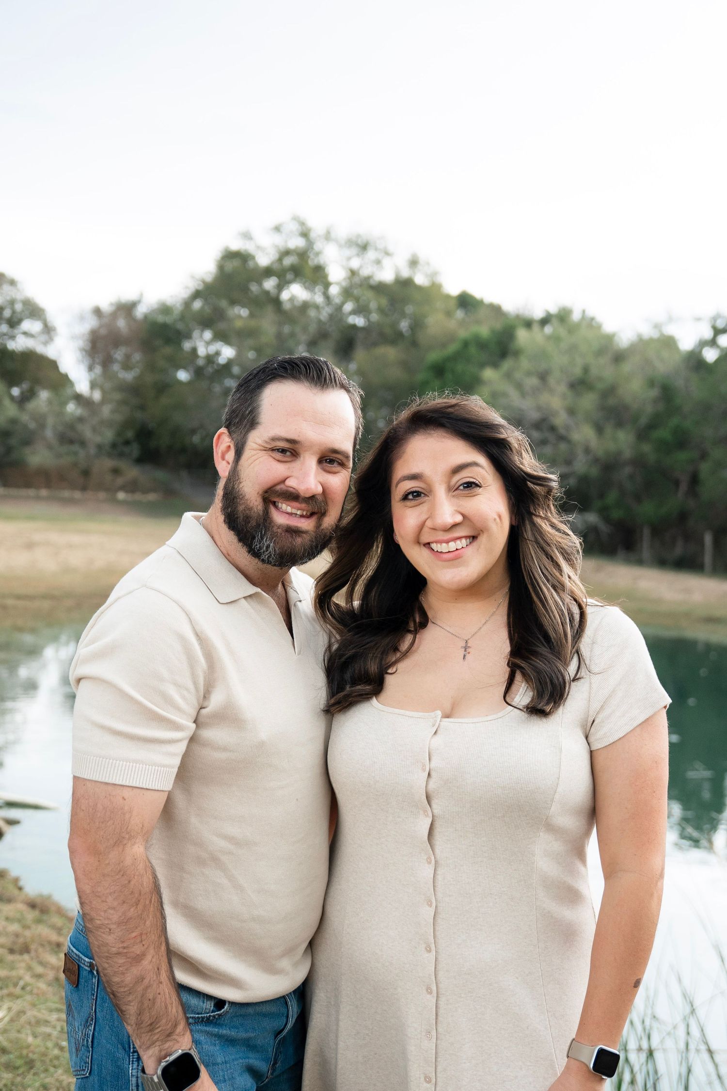 A couple wearing matching cream-colored outfits poses together by a serene lake with trees in the background.