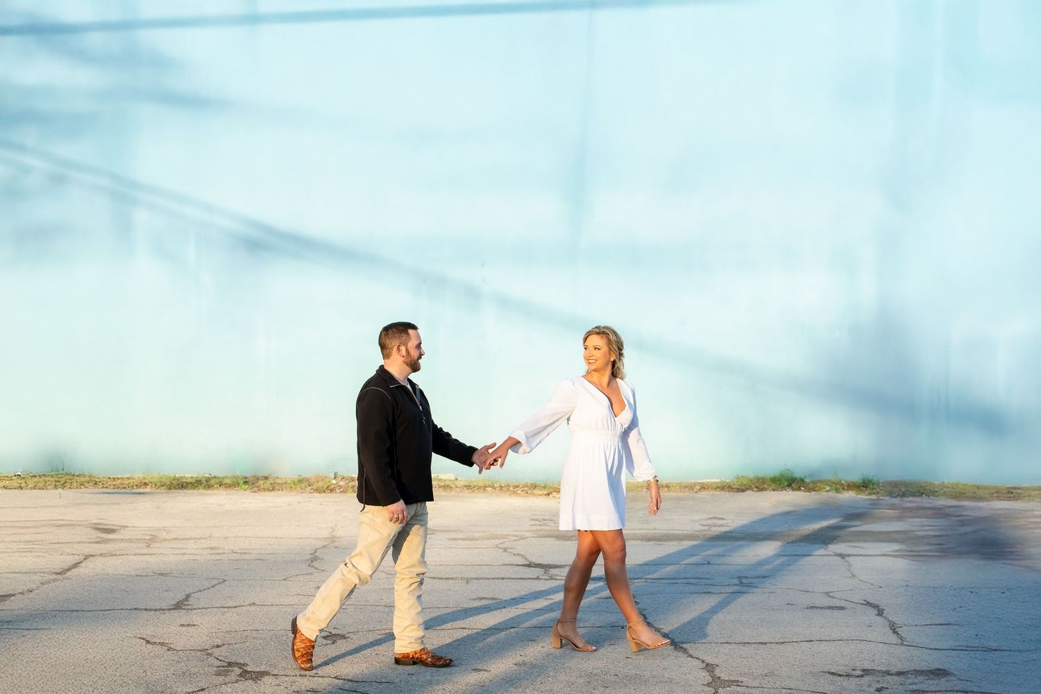 A couple in casual attire walks hand in hand on a beach during sunset with dramatic cloudy skies.