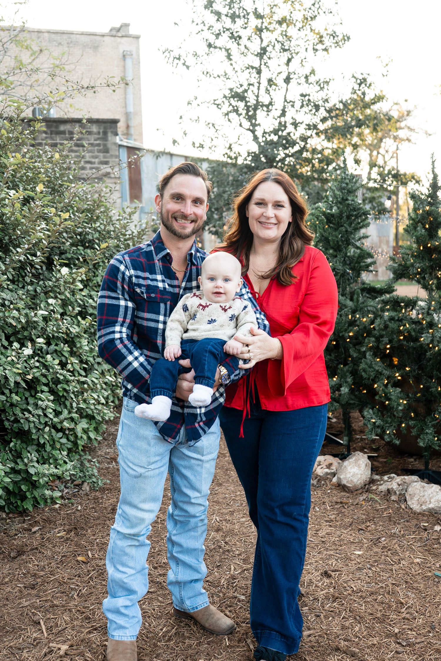 A family poses together outdoors in front of greenery wearing coordinated casual outfits of plaid and denim.