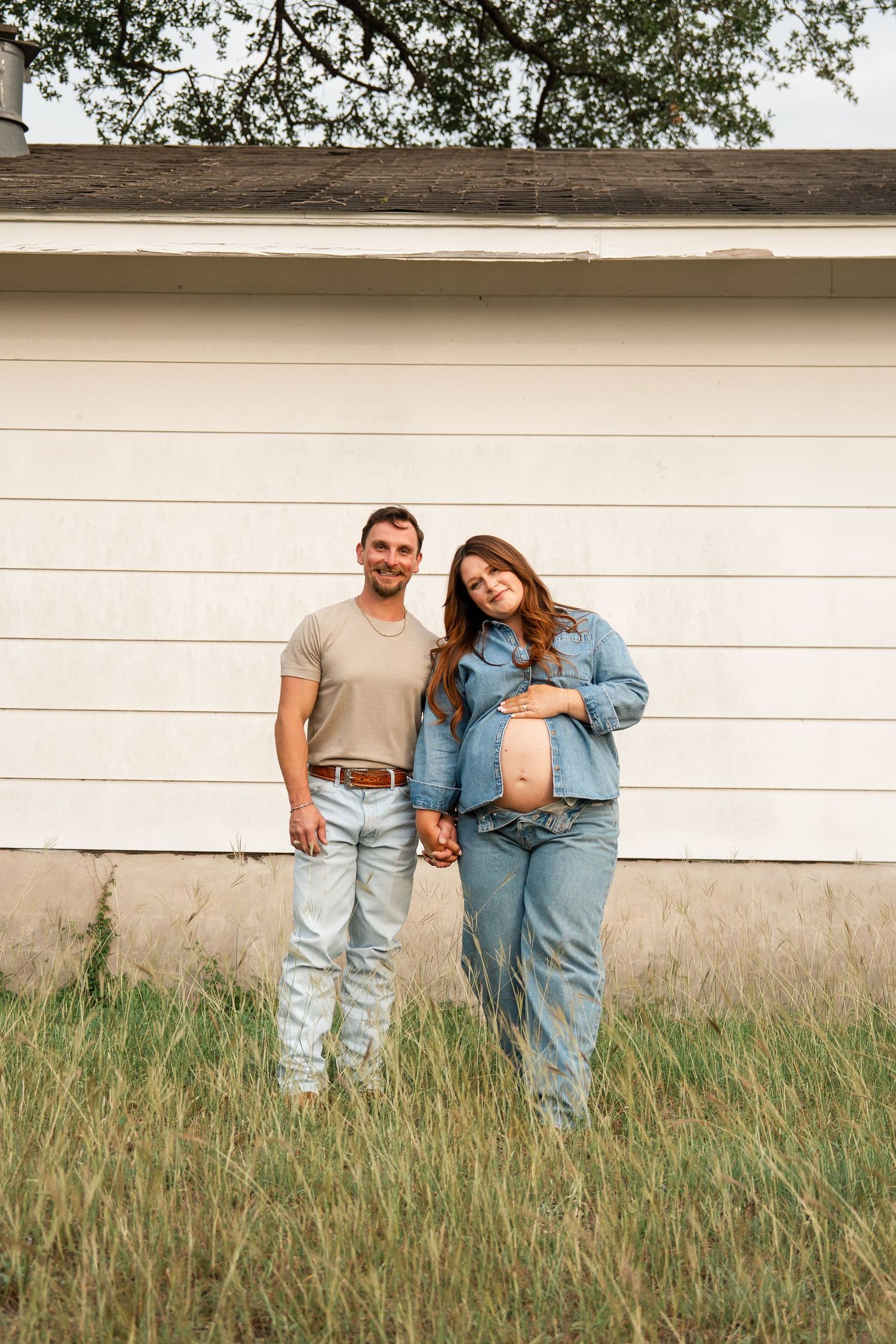 A couple in denim outfits poses casually against a white wall with tall grass in the foreground.