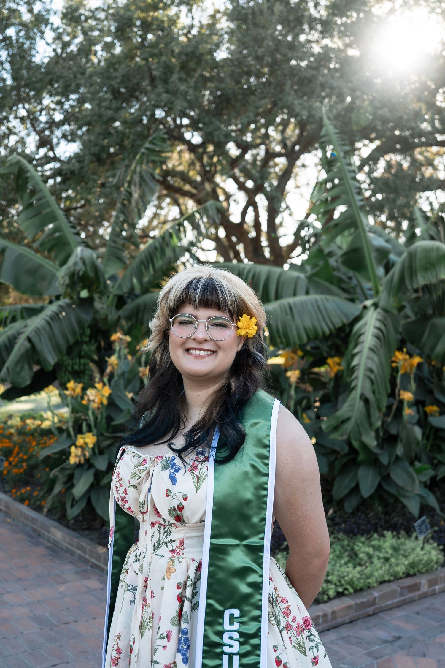 A person wearing a floral print dress poses in a lush garden setting with banana trees and flowers in the background.