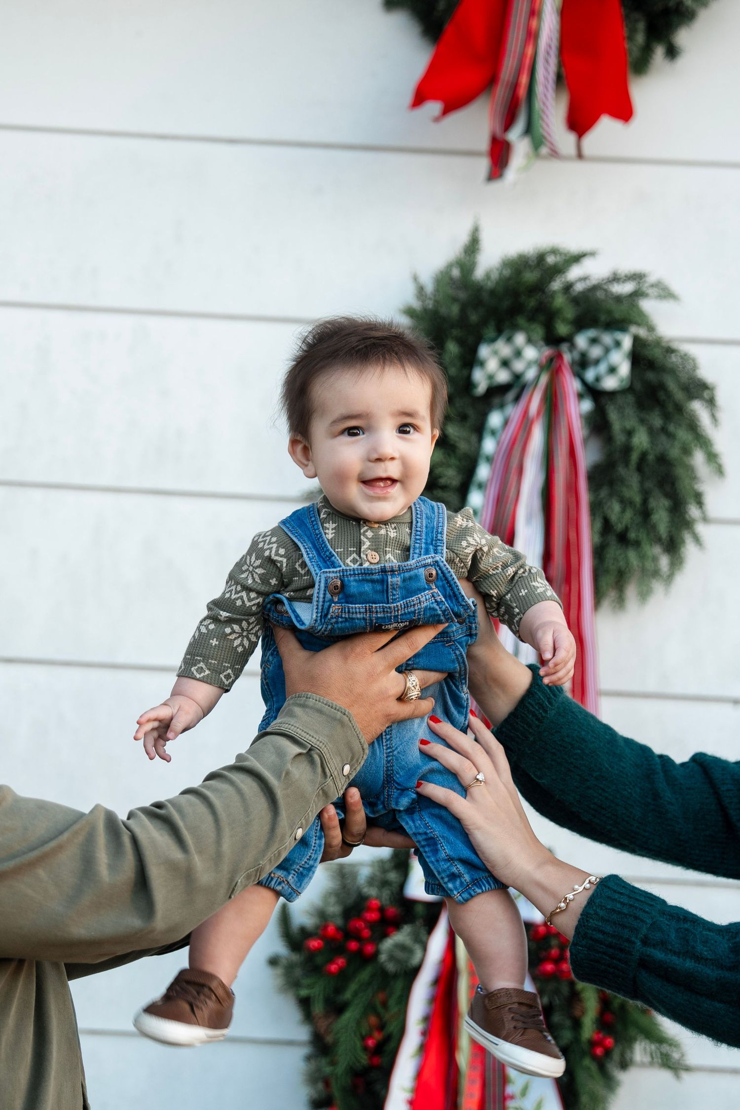 A sequence of photos showing a baby in denim overalls being lifted and held by adult hands against a holiday decorated wall.