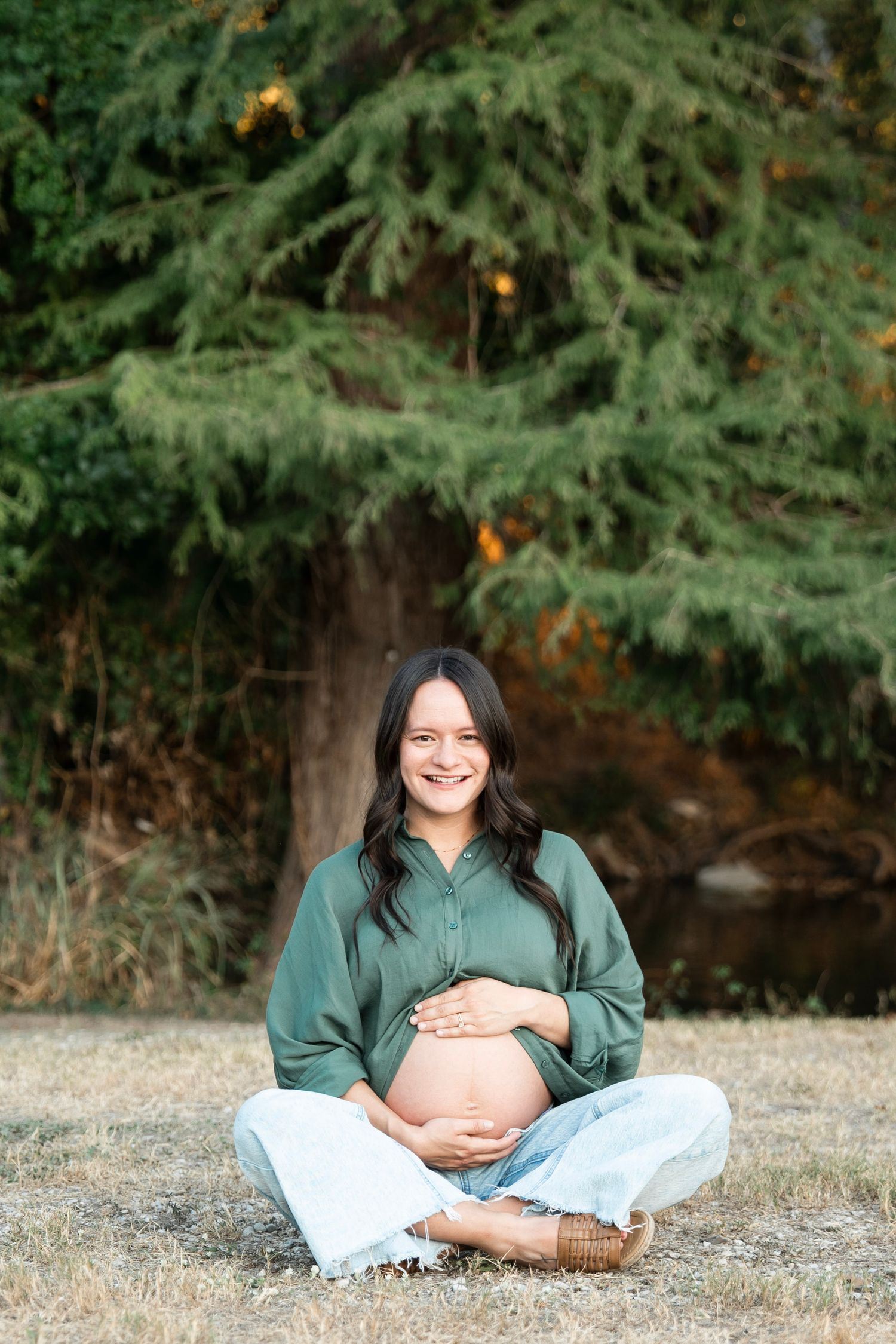 A person wearing a green sweater sits cross-legged outdoors against a backdrop of evergreen trees.