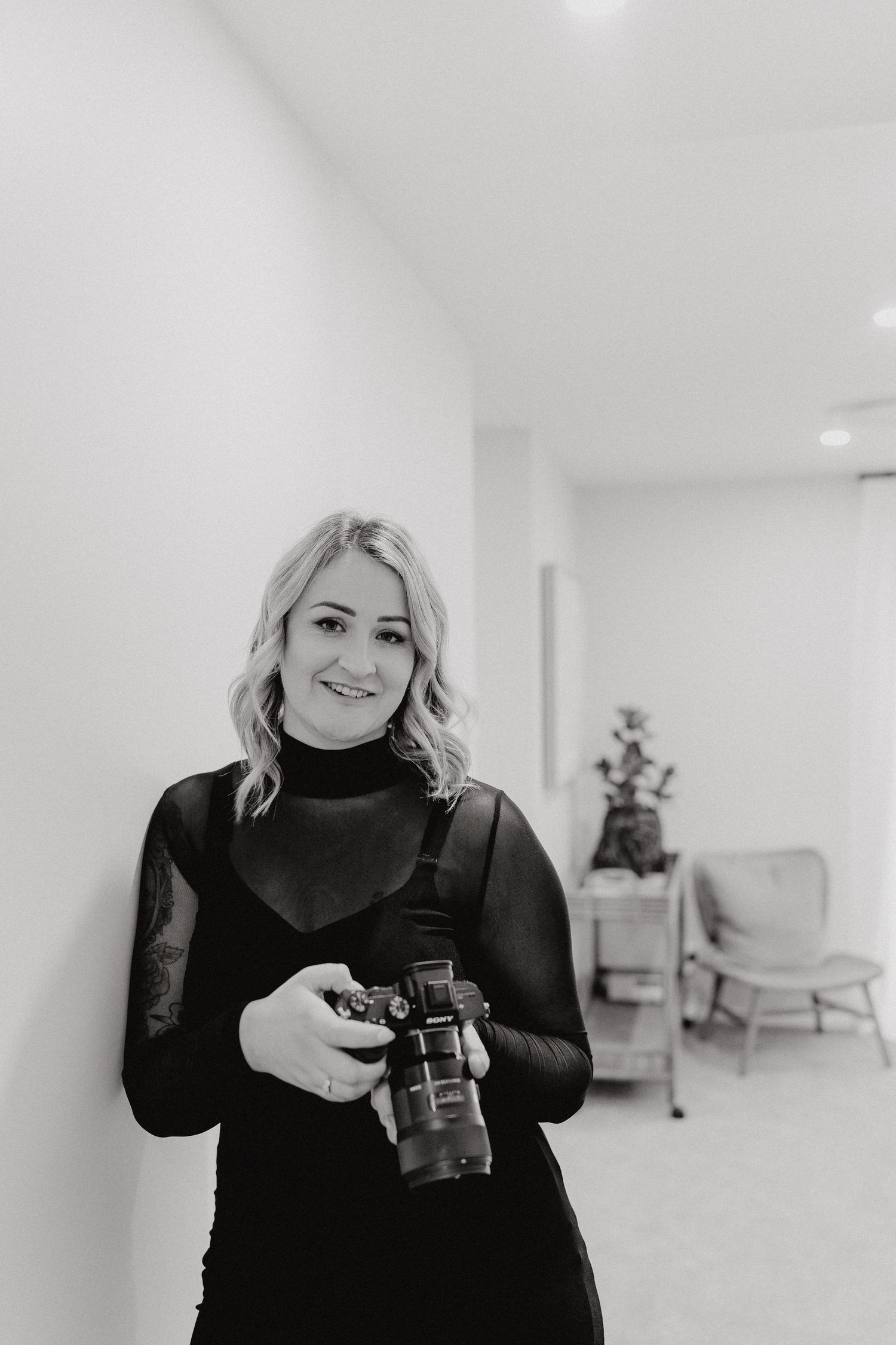A person in a black sweater holds a vintage camera while standing in a bright white minimalist room.