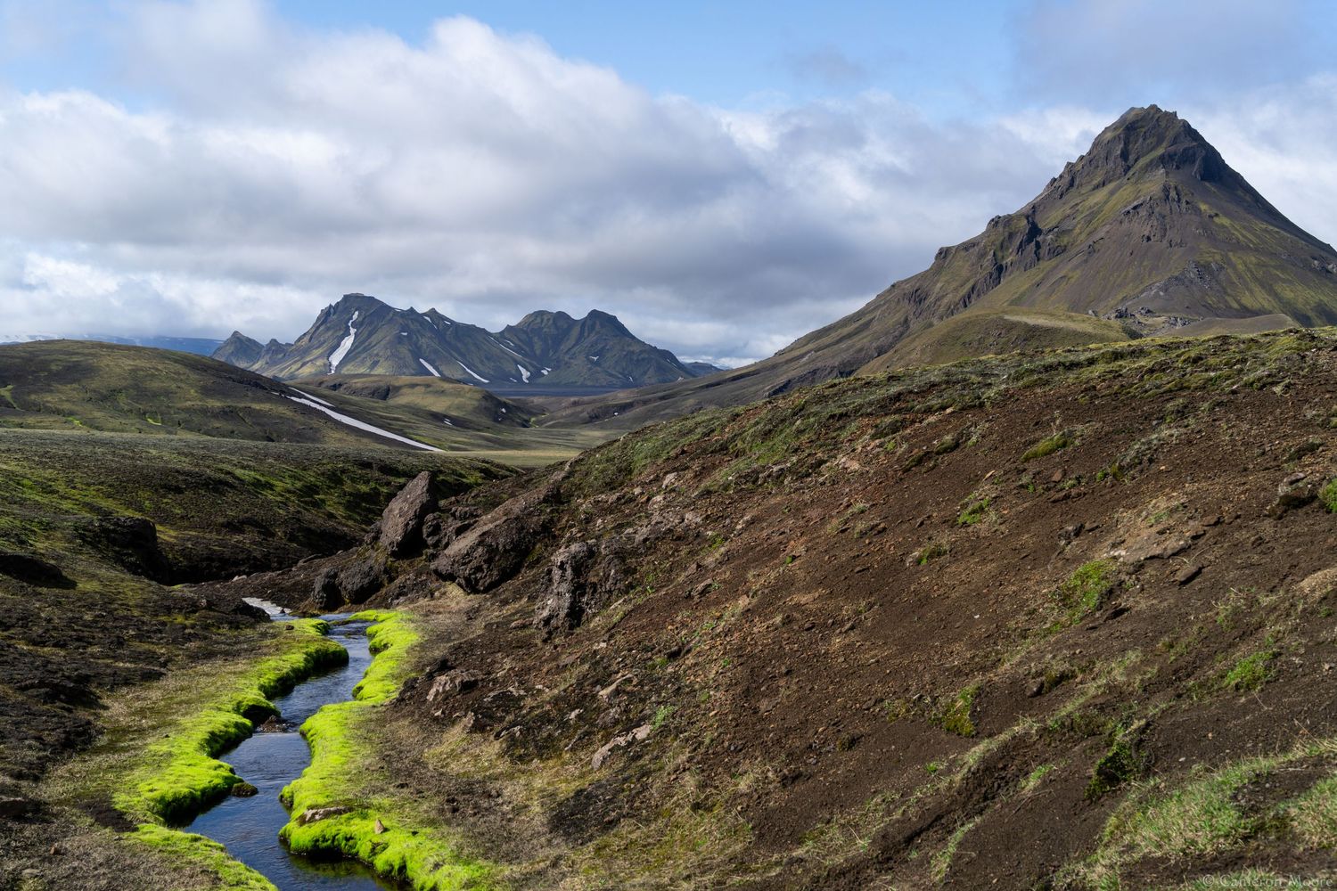 Green mossy valley with a winding stream leading toward a sharp pointed mountain peak. Iceland