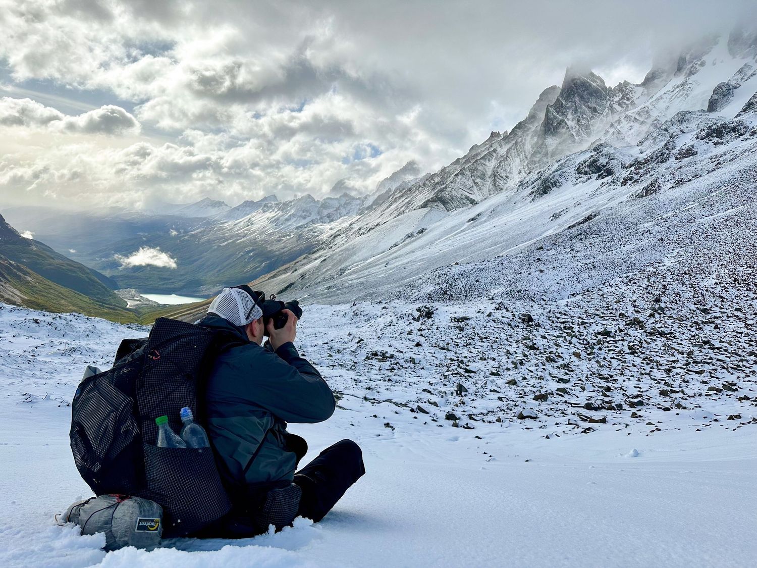 Photographer kneeling in the snow with a backpack, capturing a snowy mountain valley under a cloudy sky in Patagonia, Chile.