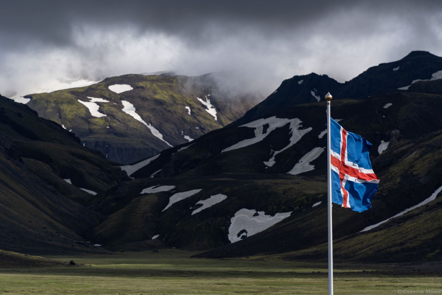 Icelandic flag flying on a pole in front of a green valley and snow-streaked mountains