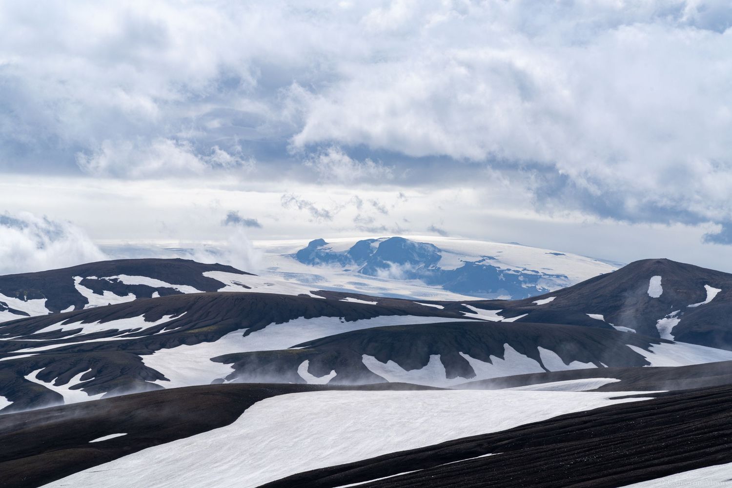 Snow-patched black volcanic mountains under a dramatic cloudy sky in a remote highland landscape. Iceland