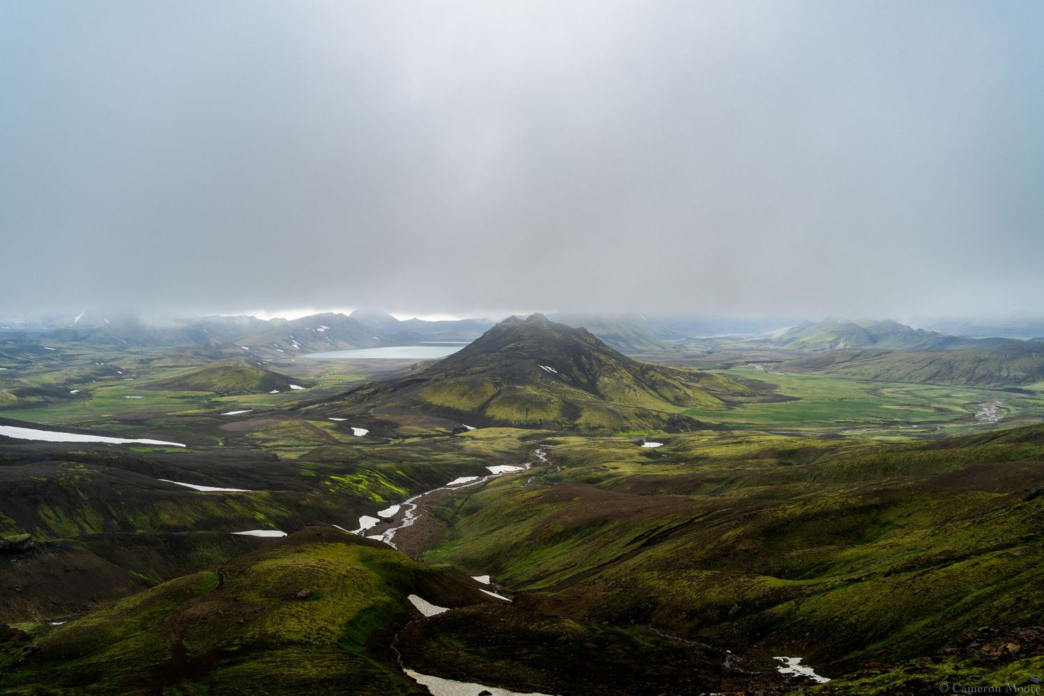 Aerial view of a lush green valley with a conical hill and a winding river beneath misty clouds. Iceland