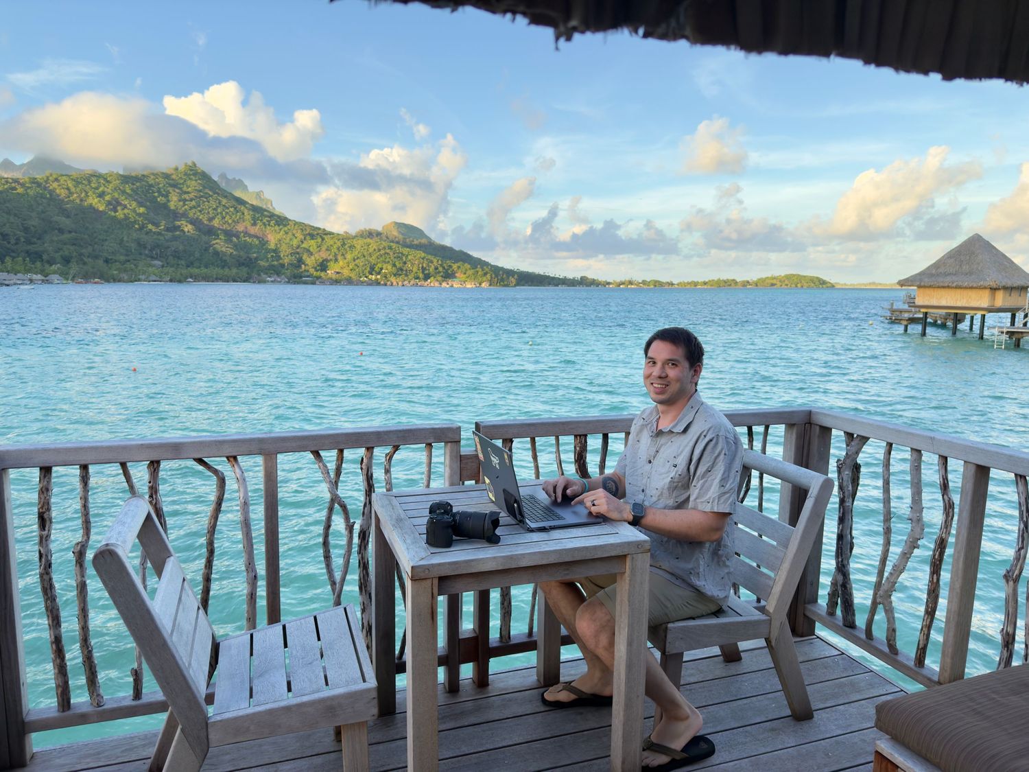 Me working on a laptop at a table on an overwater bungalow deck, turquoise lagoon and green tropical island in the background in Bora Bora.