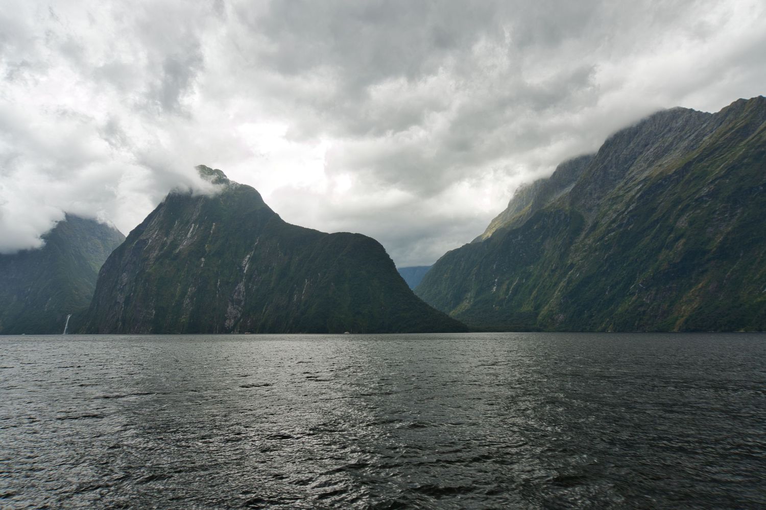 Steep mountains rising from the calm water of a fjord under heavy grey clouds at Milford Sound, New Zealand