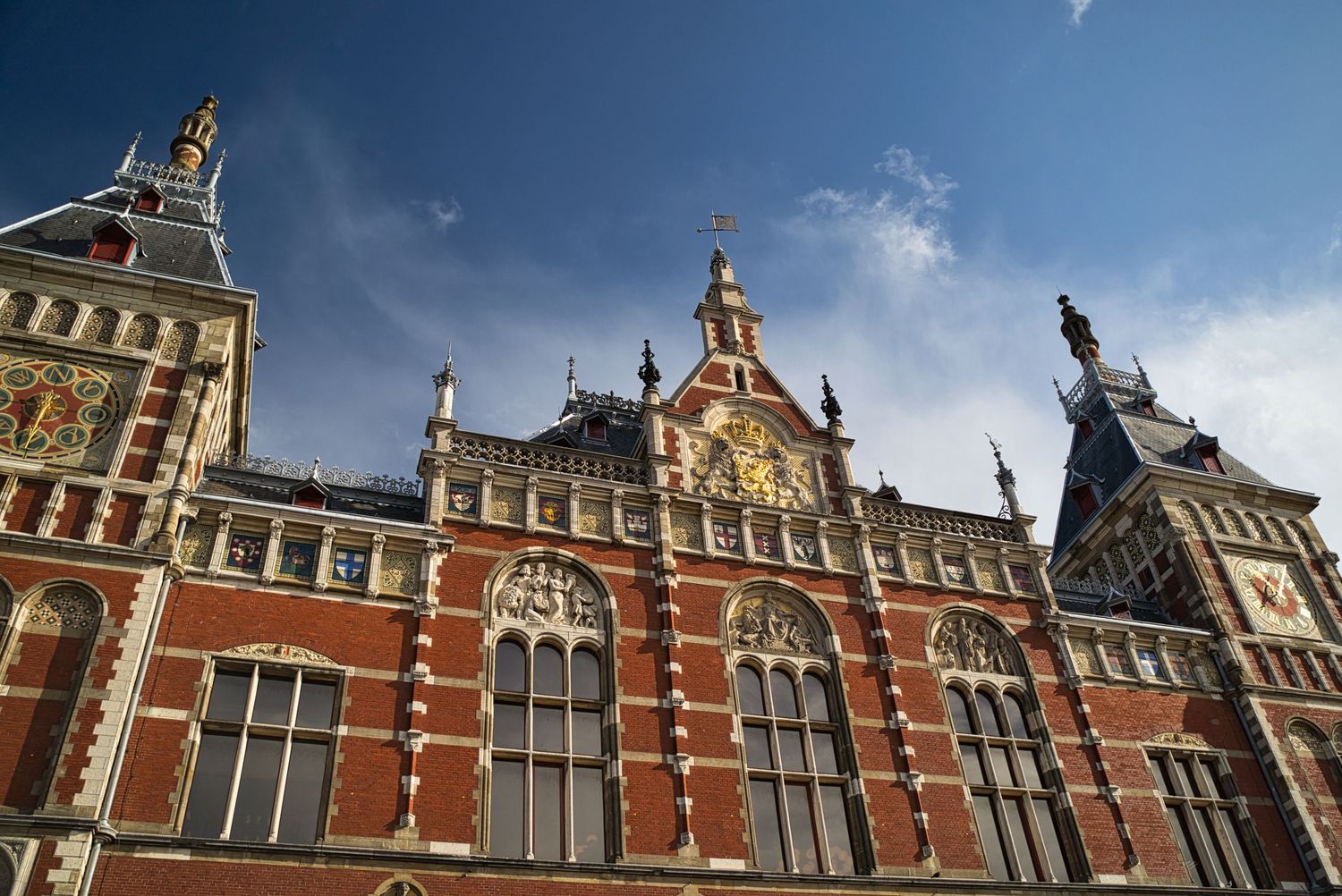 Ornate red-brick facade and clock tower of Amsterdam Central Station against a blue sky. Amsterdam