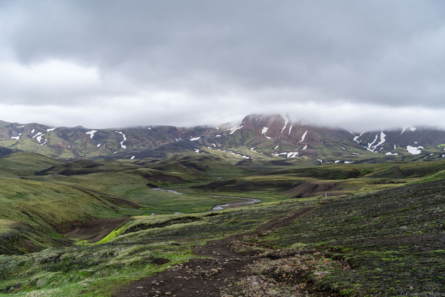Rolling green highland hills dotted with snow patches and reddish mountains under an overcast sky. Iceland