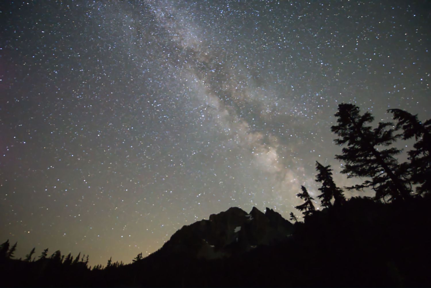 The Milky Way arching across a starry night sky above a silhouetted mountain ridge and trees. Washington