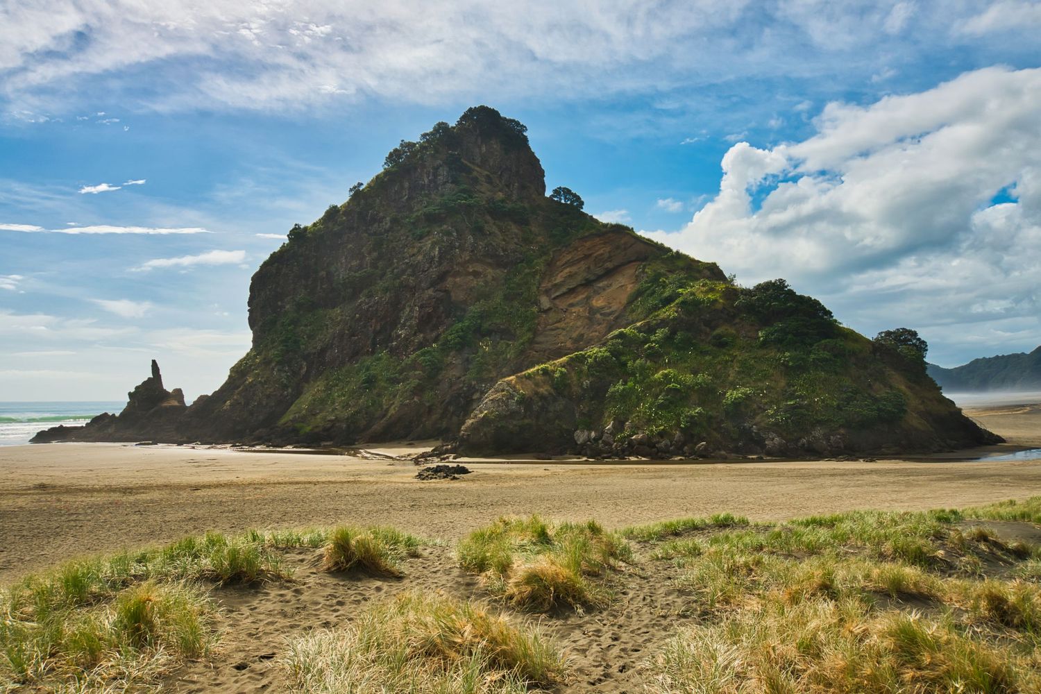 Lion Rock, a large rocky outcrop rising from a wide sandy beach under a bright blue sky. New Zealand