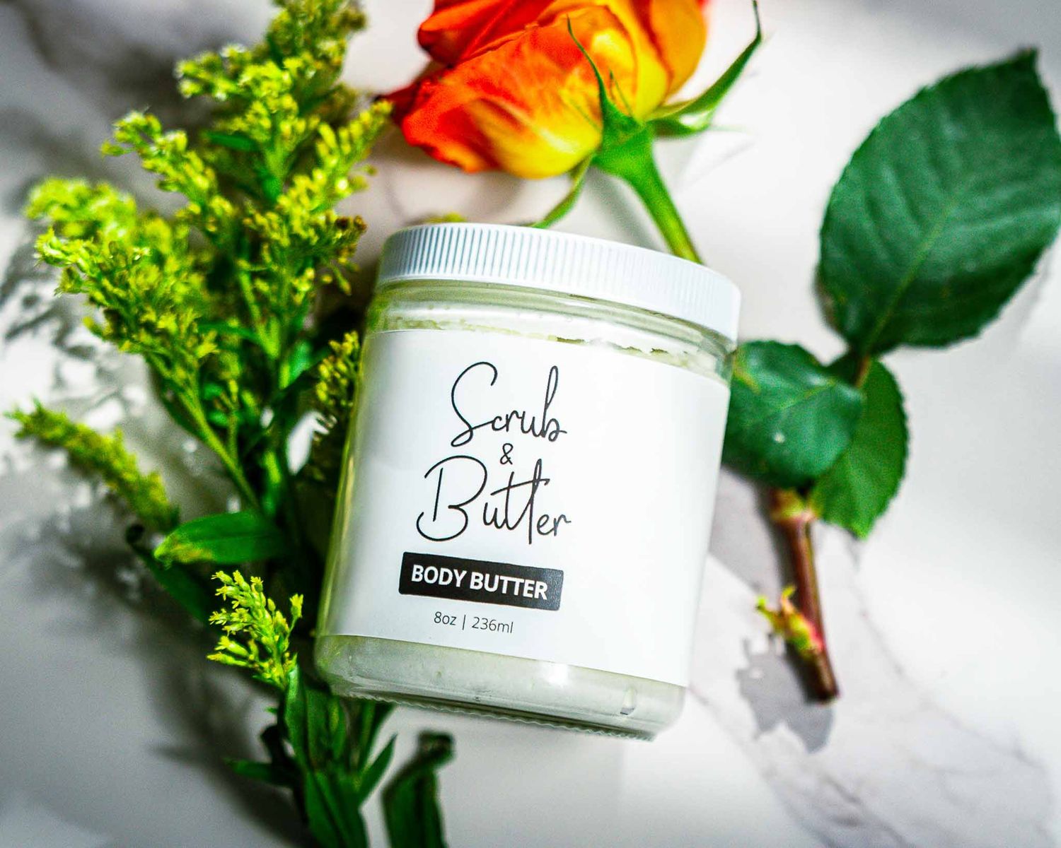 Clean product photography of body butter with orange rose and green foliage on white background.