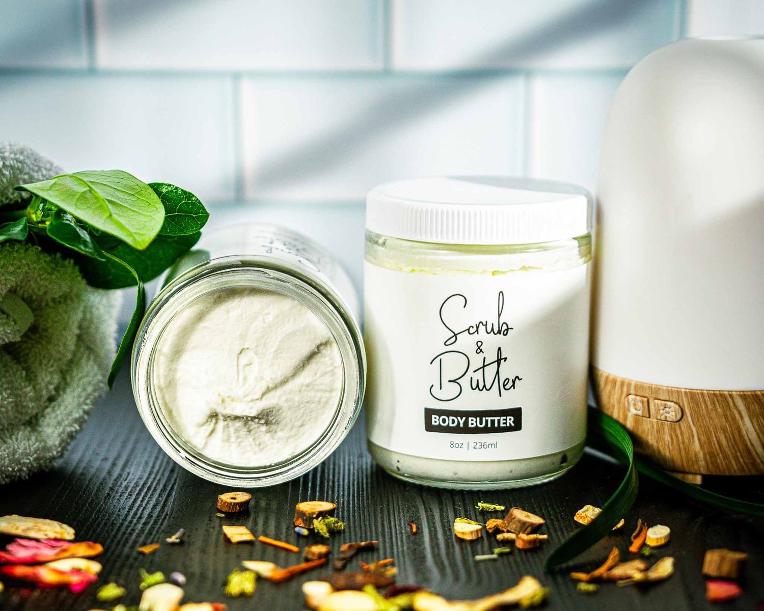 Body butter jar displayed with essential oils, diffuser and scattered flower petals on wooden surface.