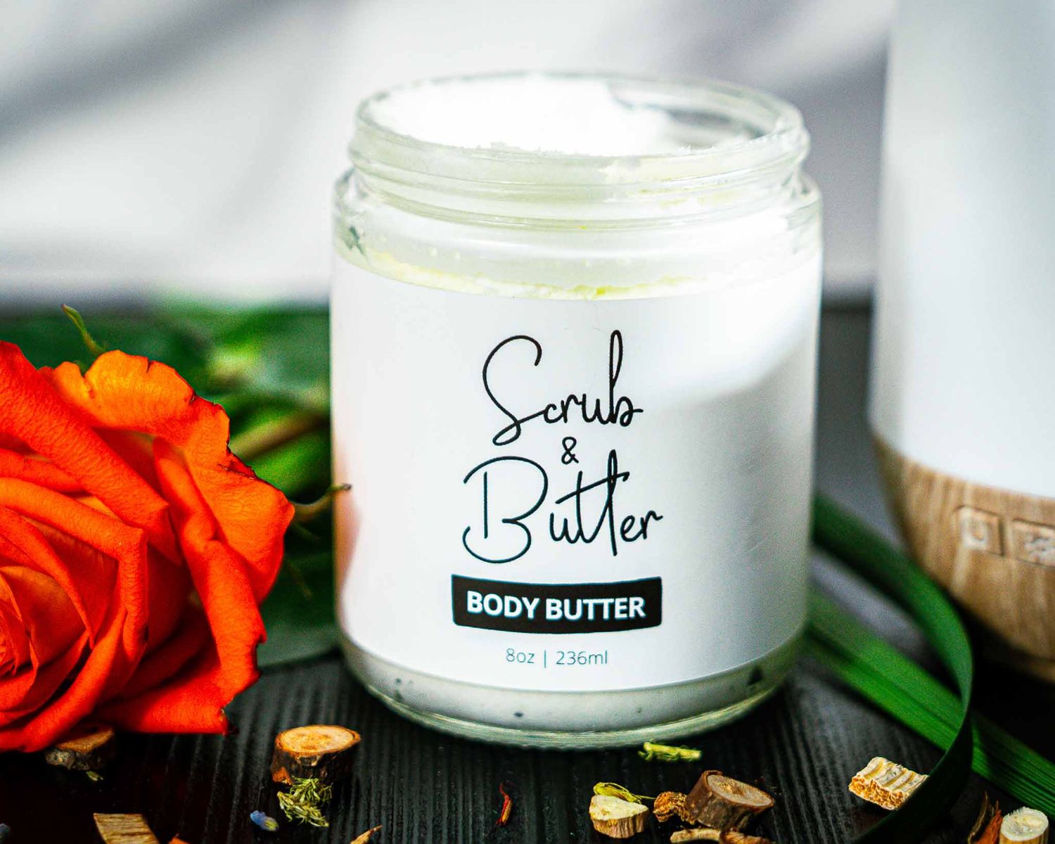 Body butter product jar photographed with vibrant orange rose and scattered nuts on wooden surface.