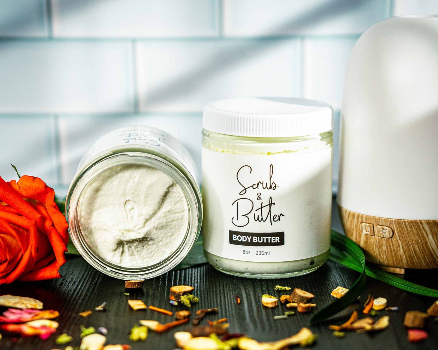 Two jars of body butter displayed with orange rose petals and wooden diffuser in soft lighting.