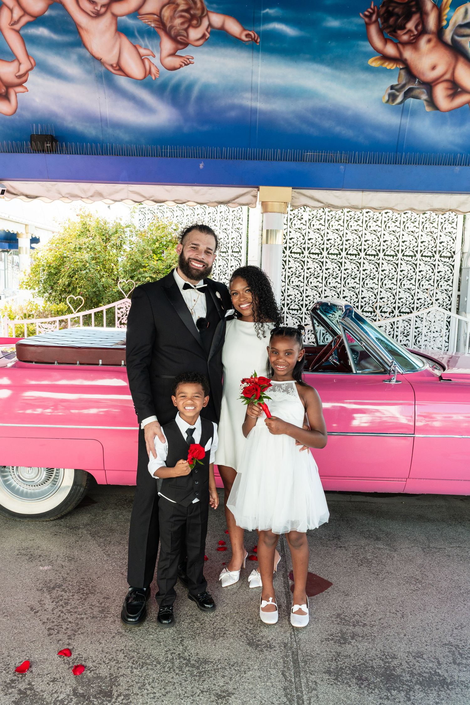 Wedding party poses together next to a pink vintage car under a blue canopy.
