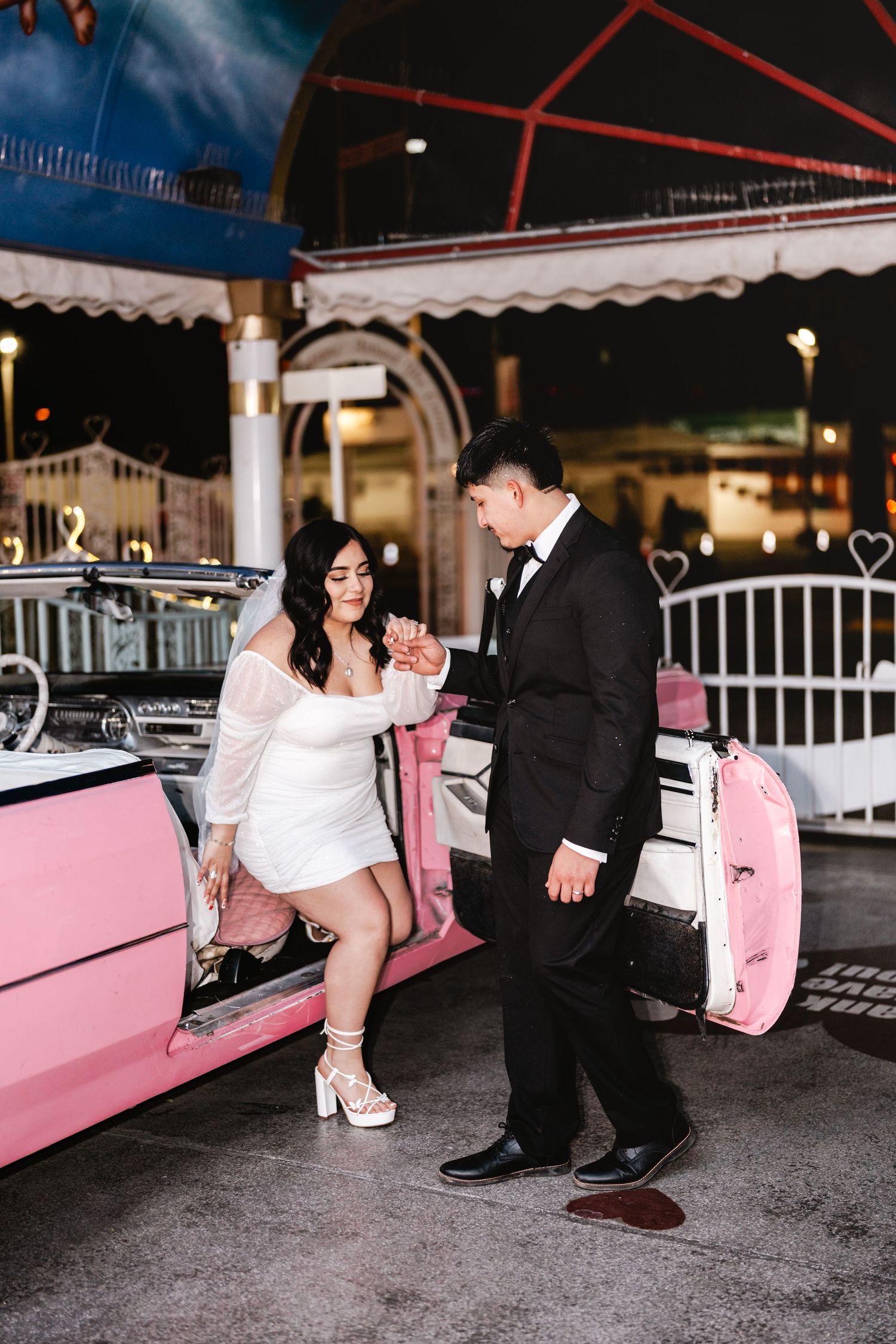 A romantic couple poses beside a pink classic car in a vintage-style wedding photoshoot.