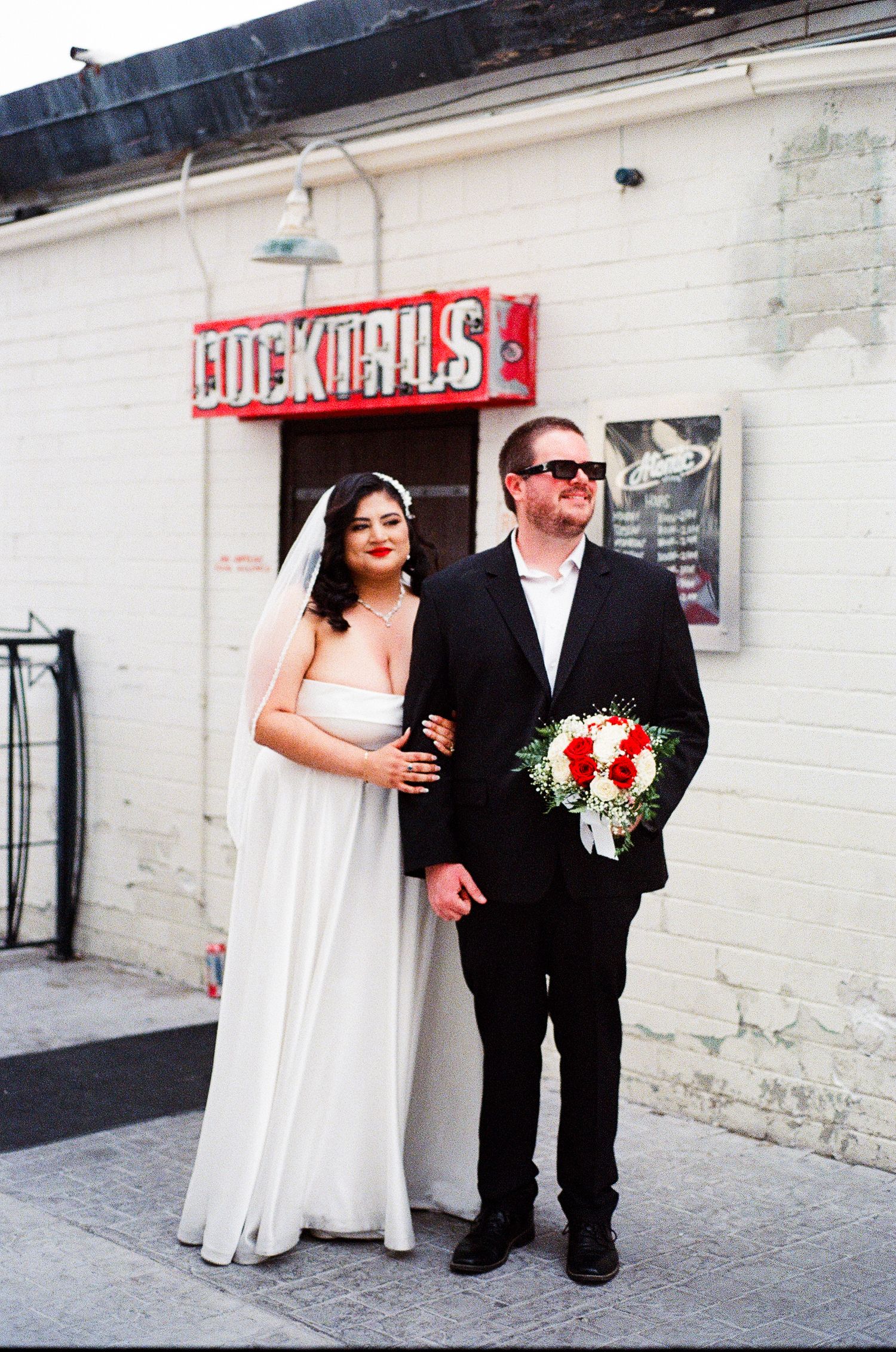 A couple poses outside a venue with a red neon sign and white brick wall.