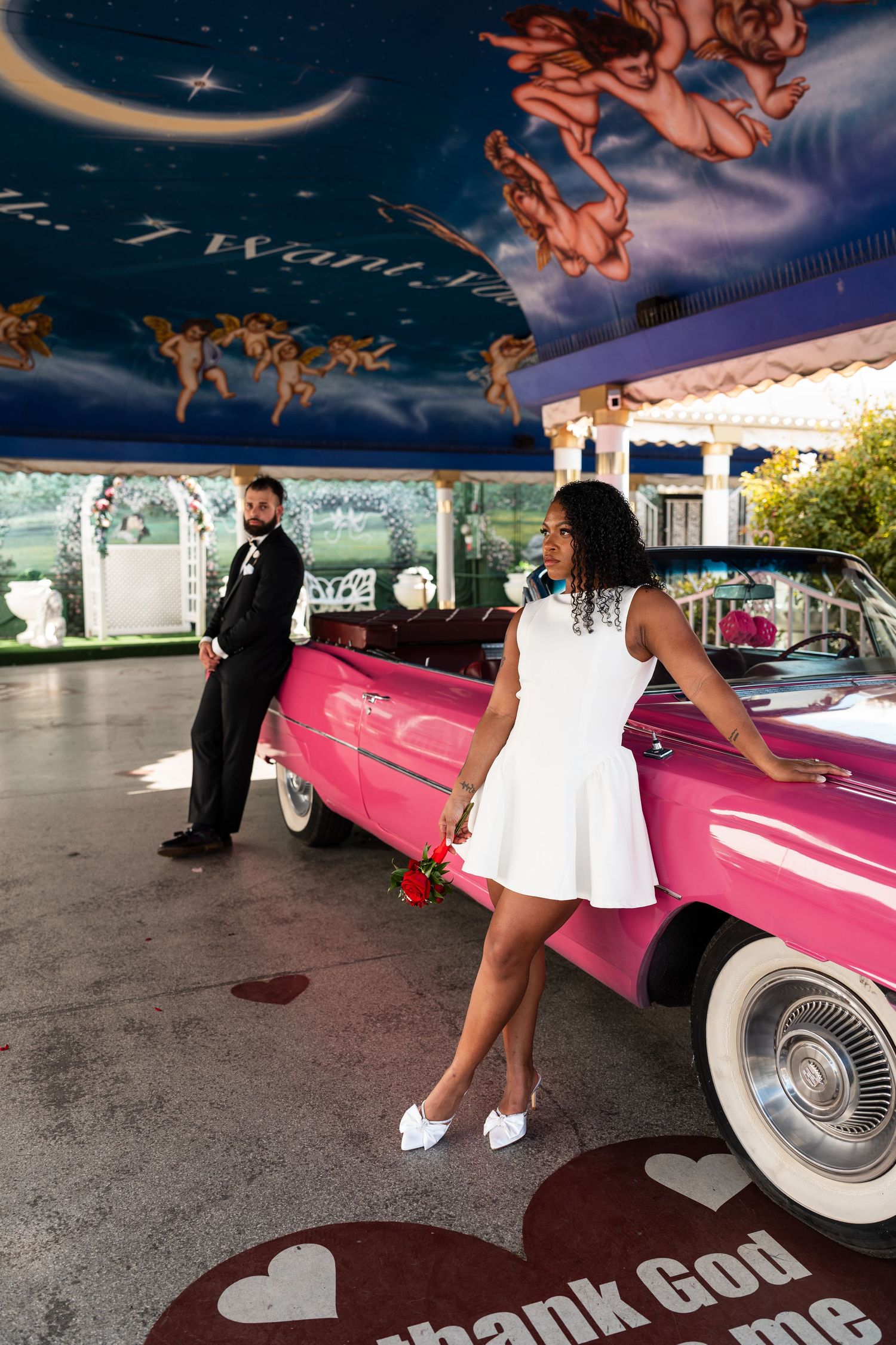 Wedding couple poses for romantic photos with a pink classic car under blue skies.
