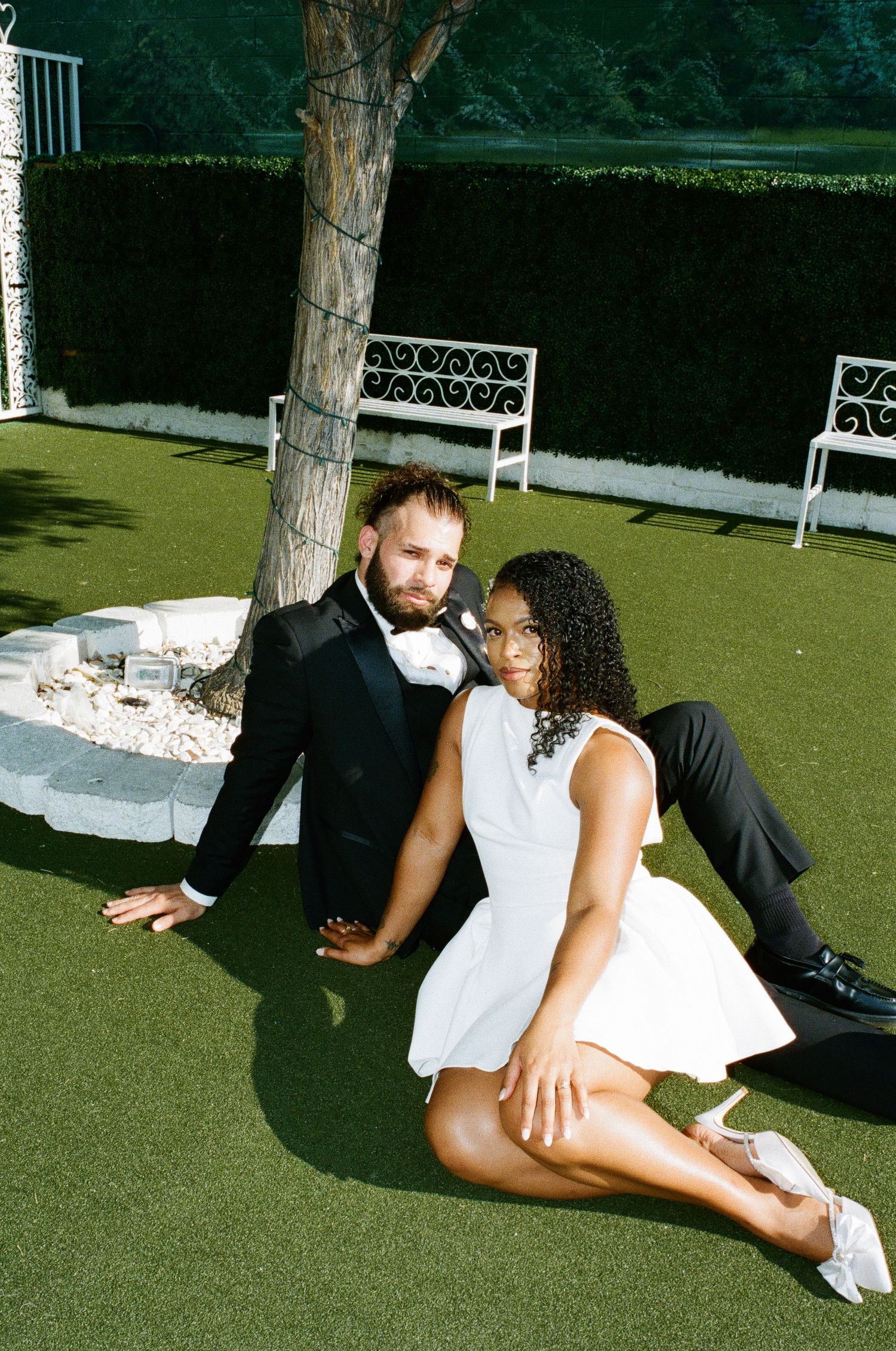 A couple relaxes on artificial turf with white garden benches in the background.