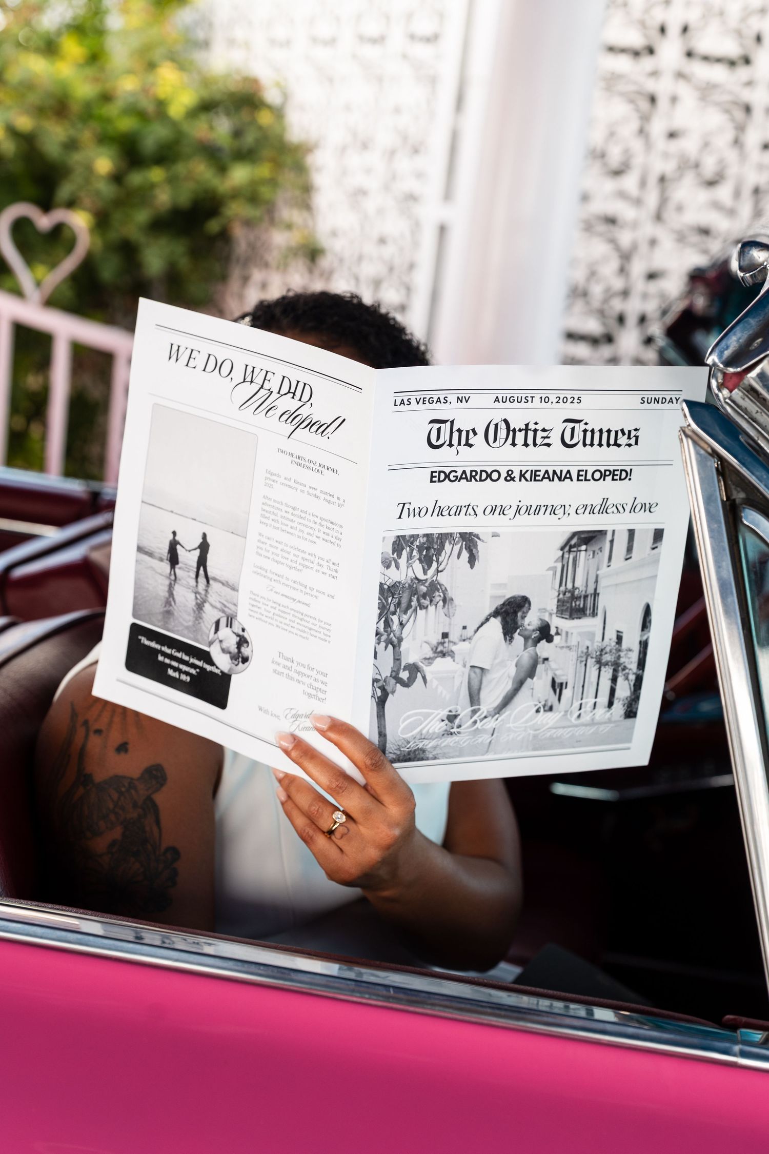 Wedding couple reading newspaper spreads in a classic pink car.