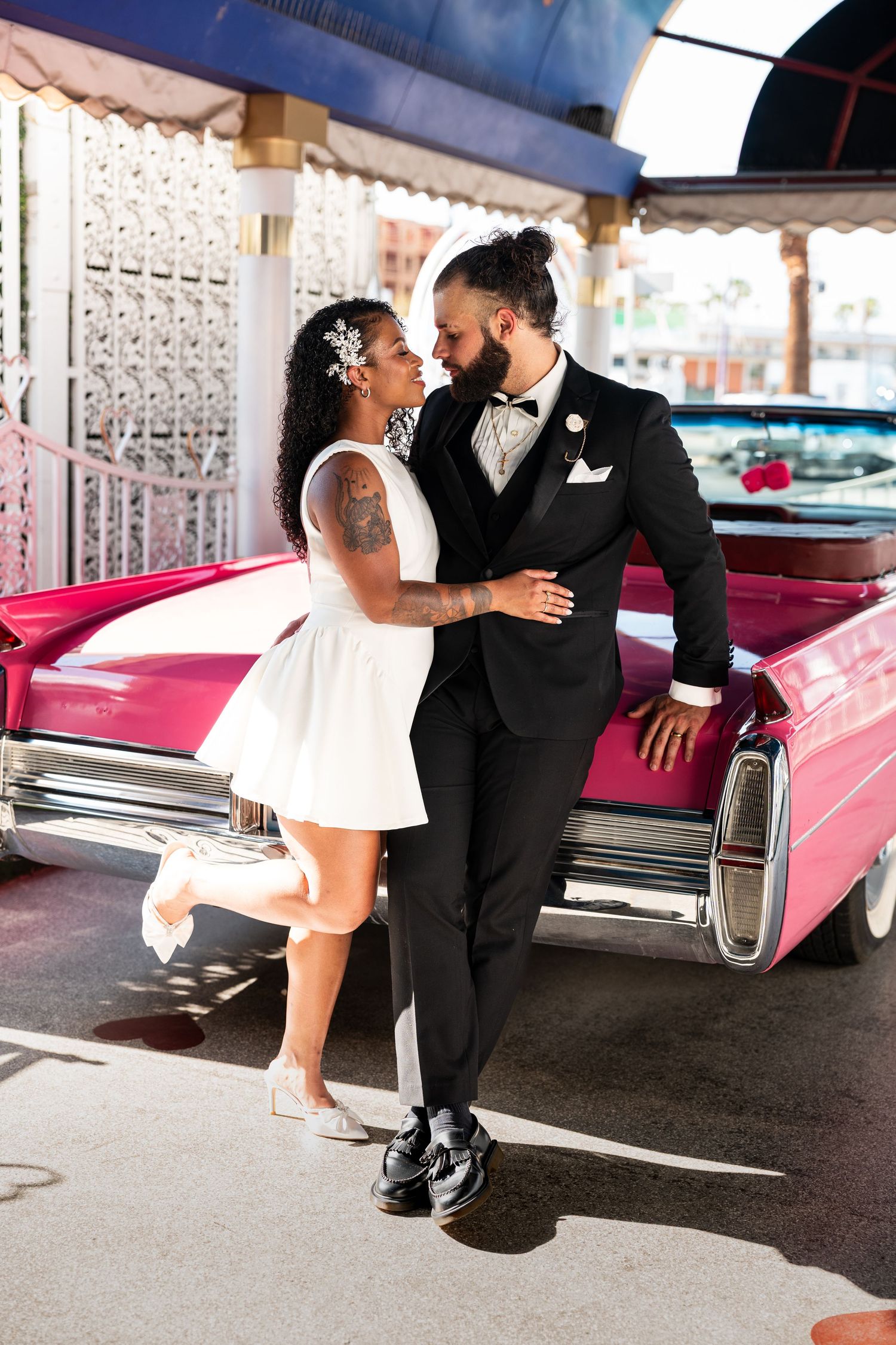 A couple poses romantically against a pink classic car at a retro-style venue.