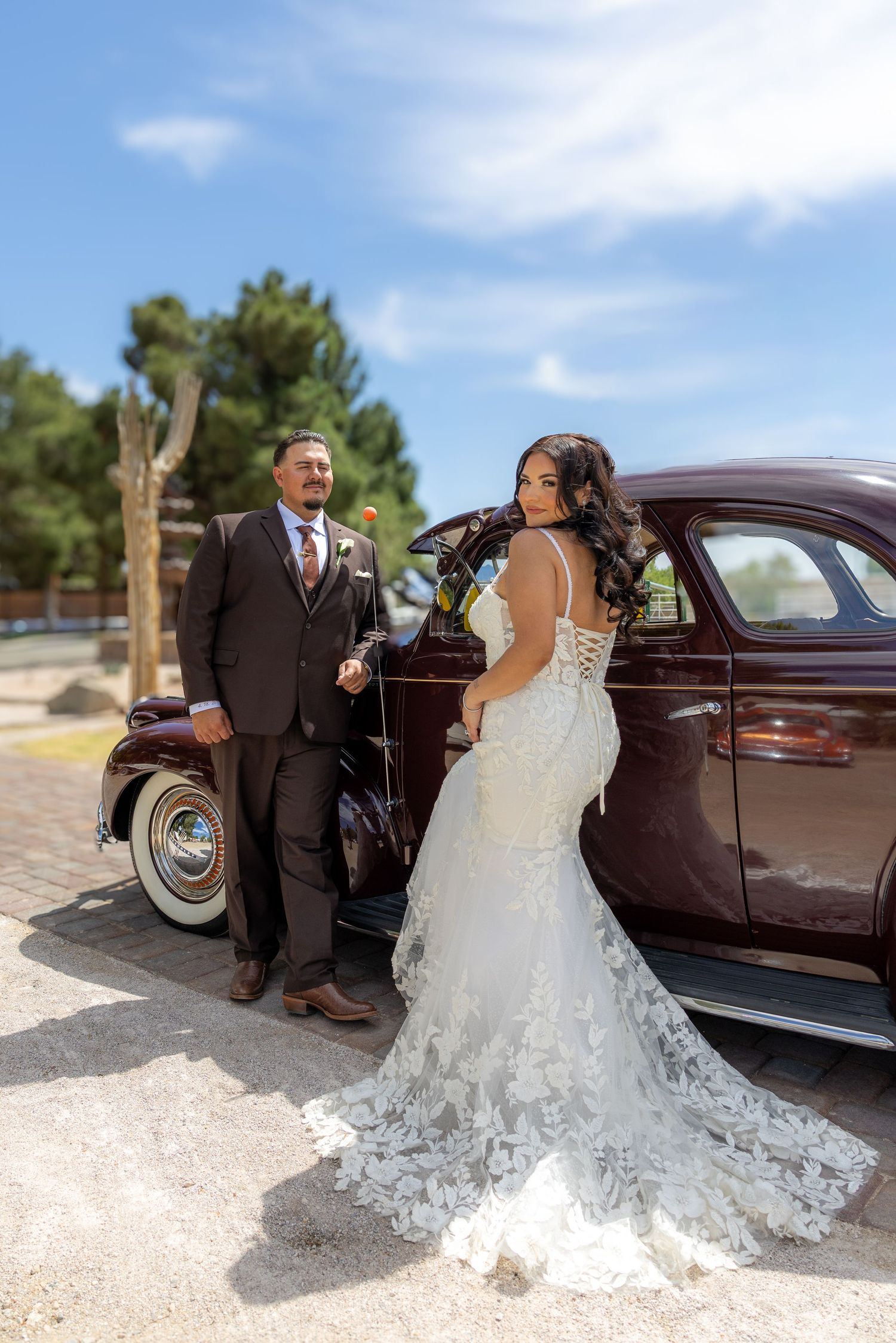 Bride in lace mermaid gown and groom in brown suit pose beside a vintage maroon car outdoors on a sunny day.