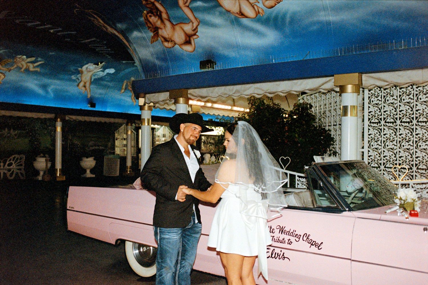 A couple celebrates near a vintage pink Cadillac at a retro-themed venue.