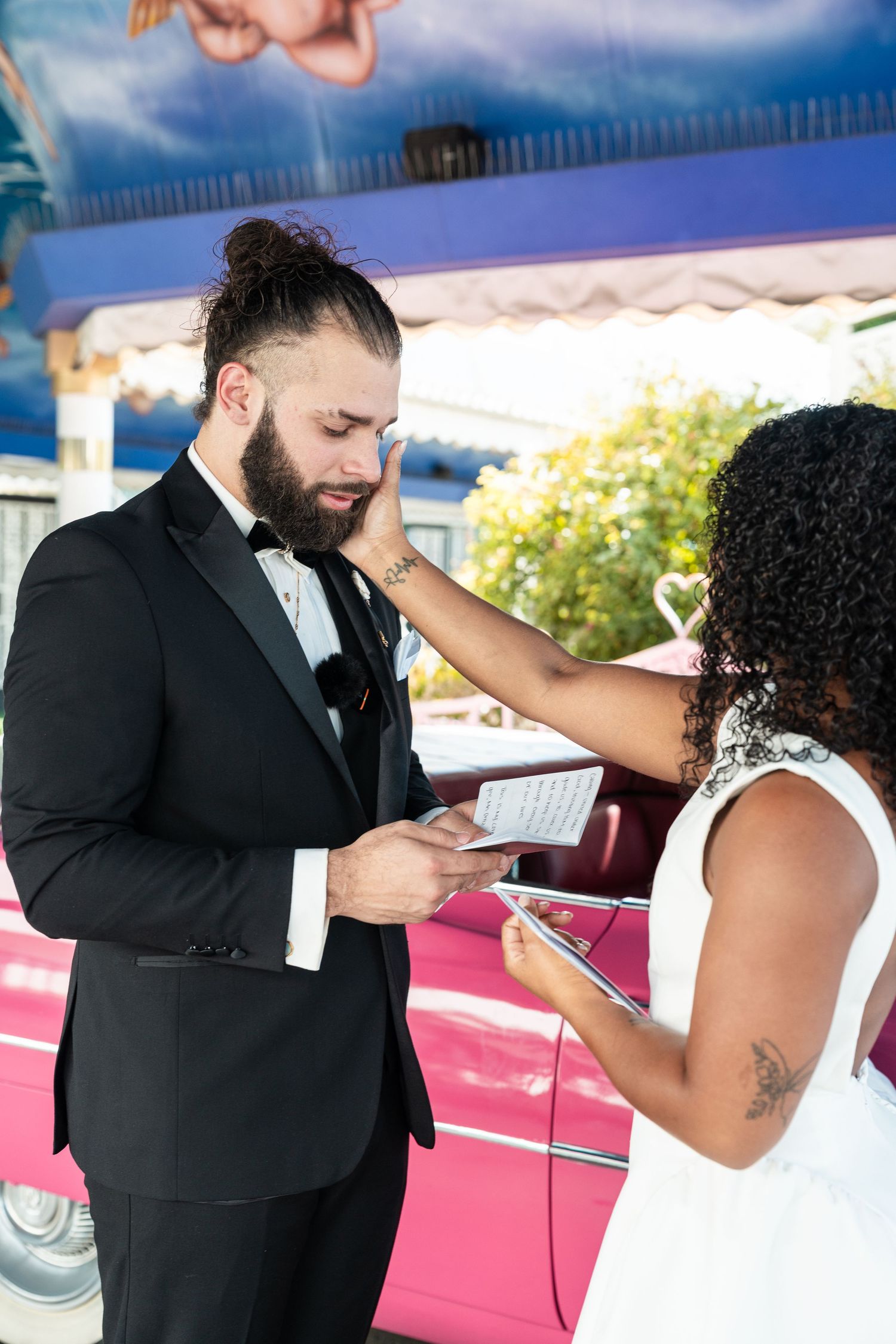 Newlyweds share a tender moment beside a pink classic car during their wedding.