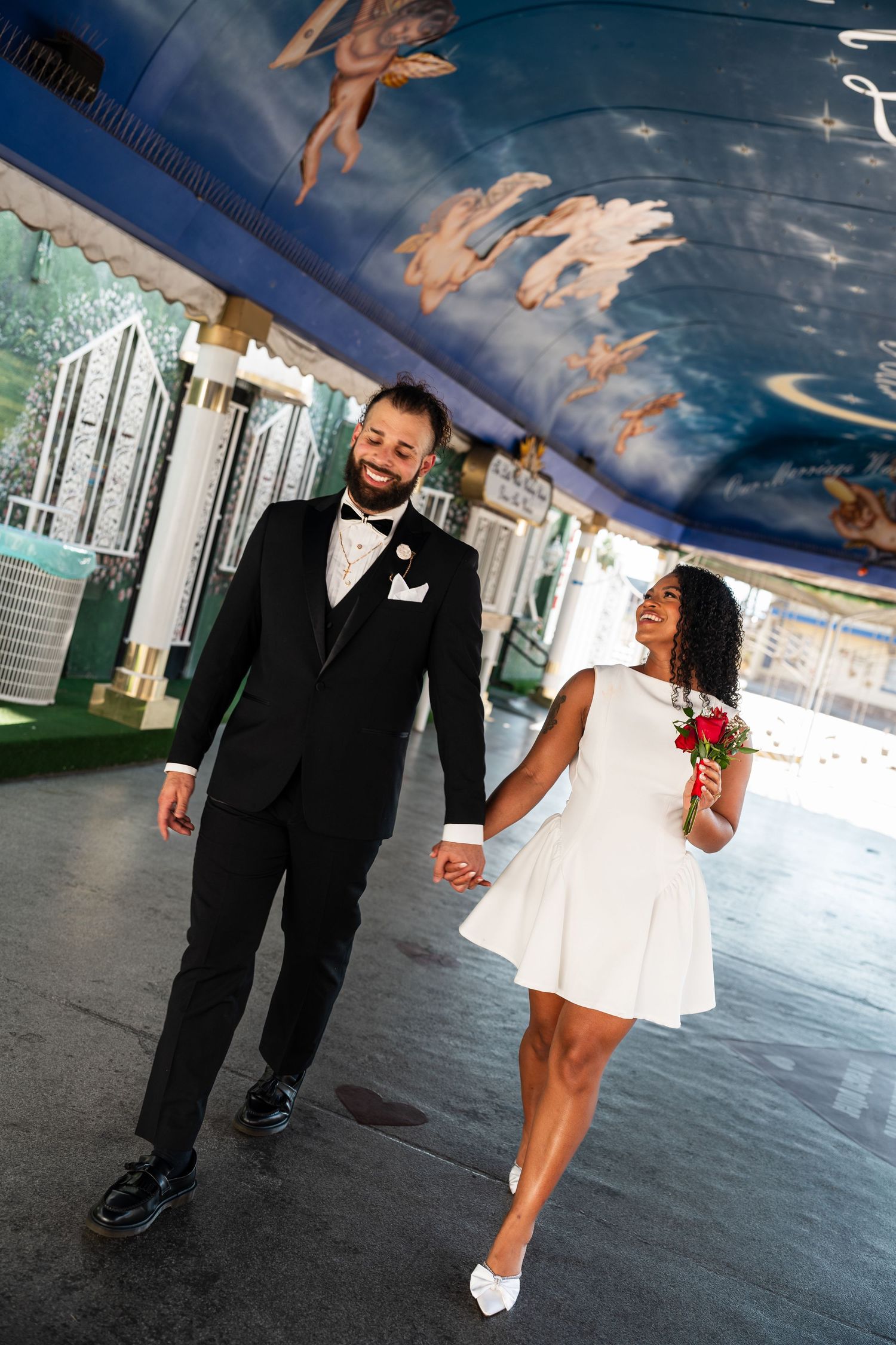 A wedding couple walks beneath a decorated tunnel with painted cherubs on the ceiling.