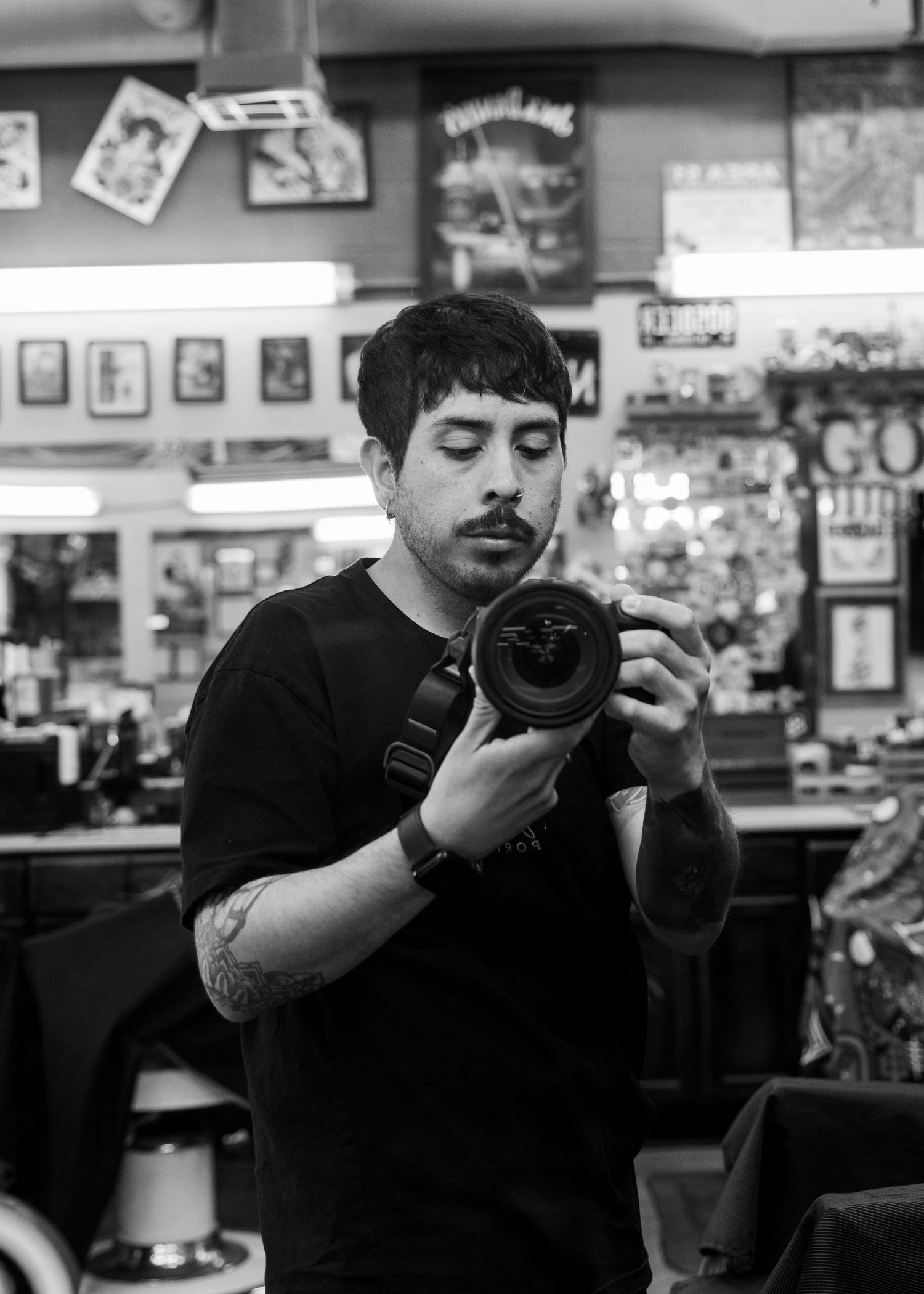 Black and white photo of a young tattooed man taking a mirror selfie with a DSLR camera in a tattoo shop.