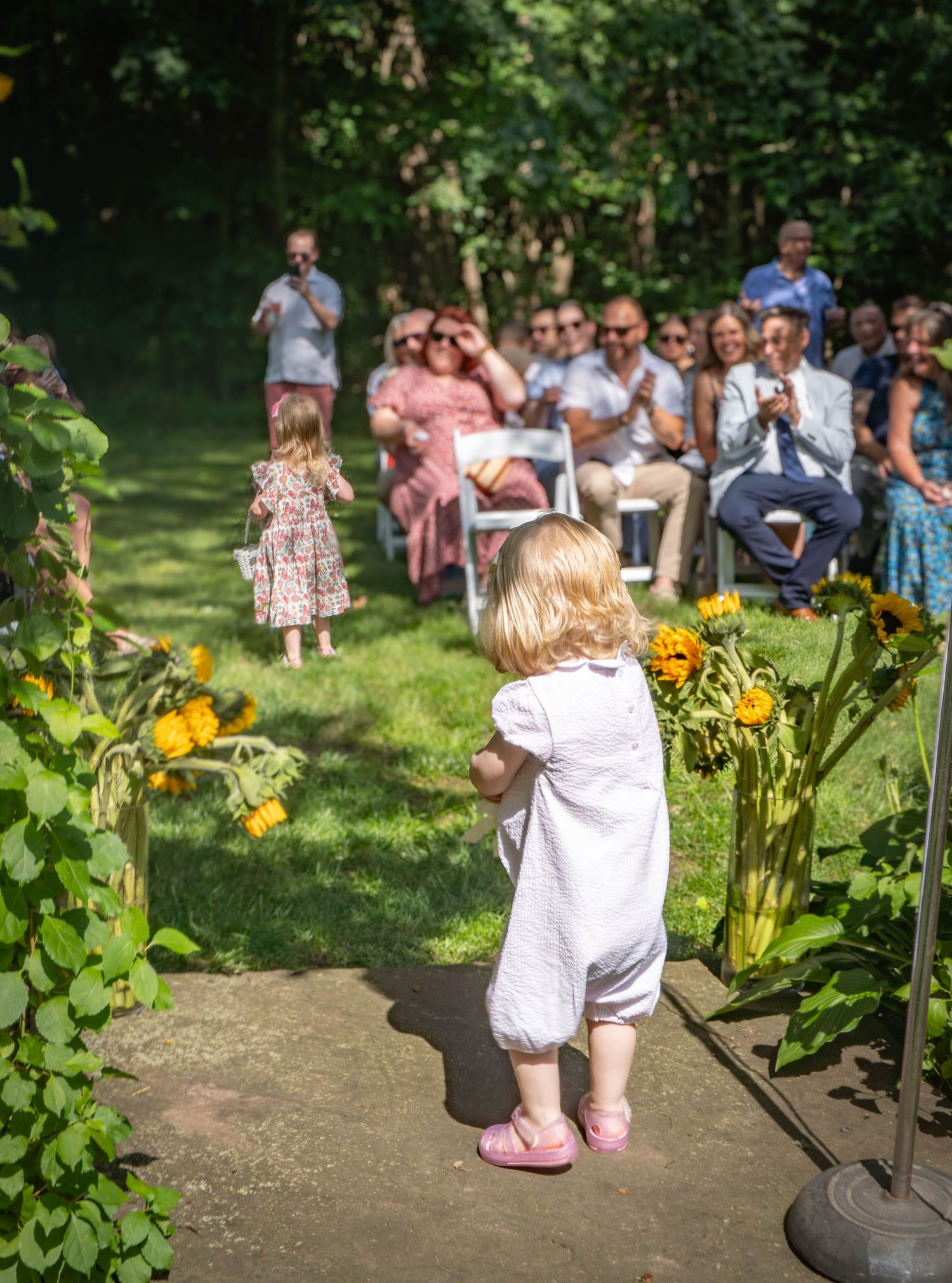 Two toddler flower girls walking down a sunflower-lined outdoor wedding aisle toward seated guests.