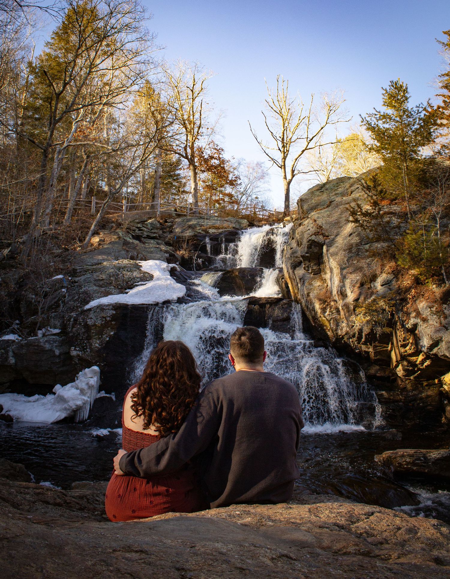 Couple sitting together on rocks admiring a cascading waterfall surrounded by autumn trees.