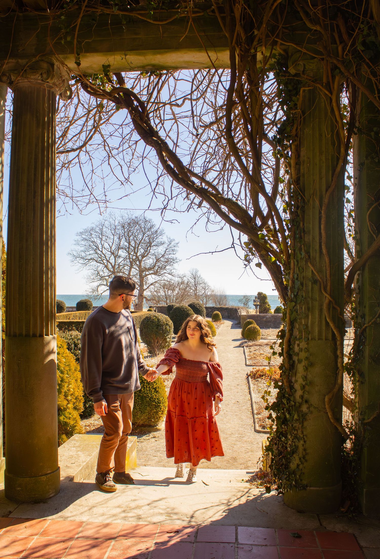 Couple holding hands walking through sunlit stone pergola in a formal winter garden.