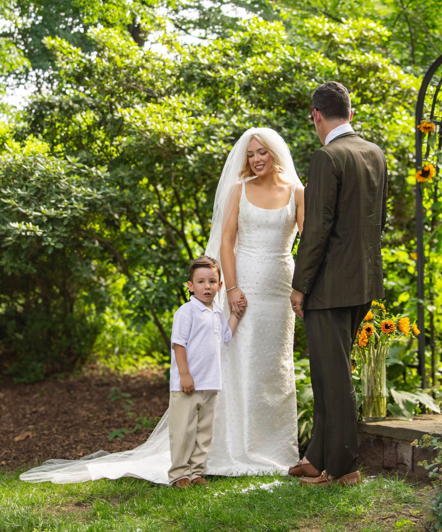 Smiling bride in white gown holding hands with young boy at outdoor wedding ceremony.