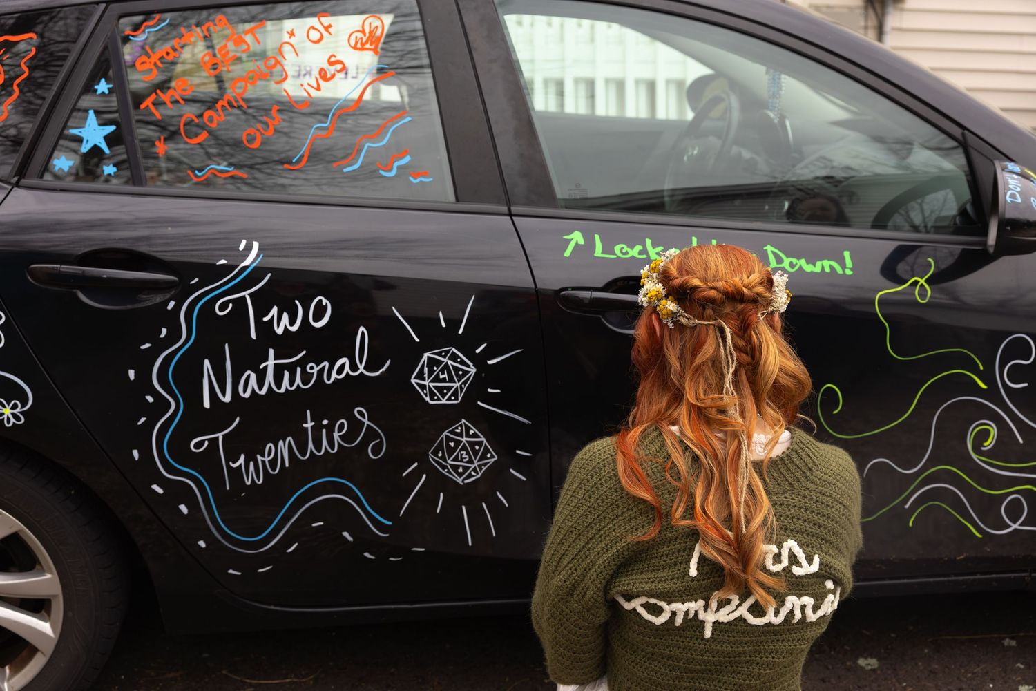 Bride in green sweater reads chalk art decorations on black car reading Two Natural Twenties.