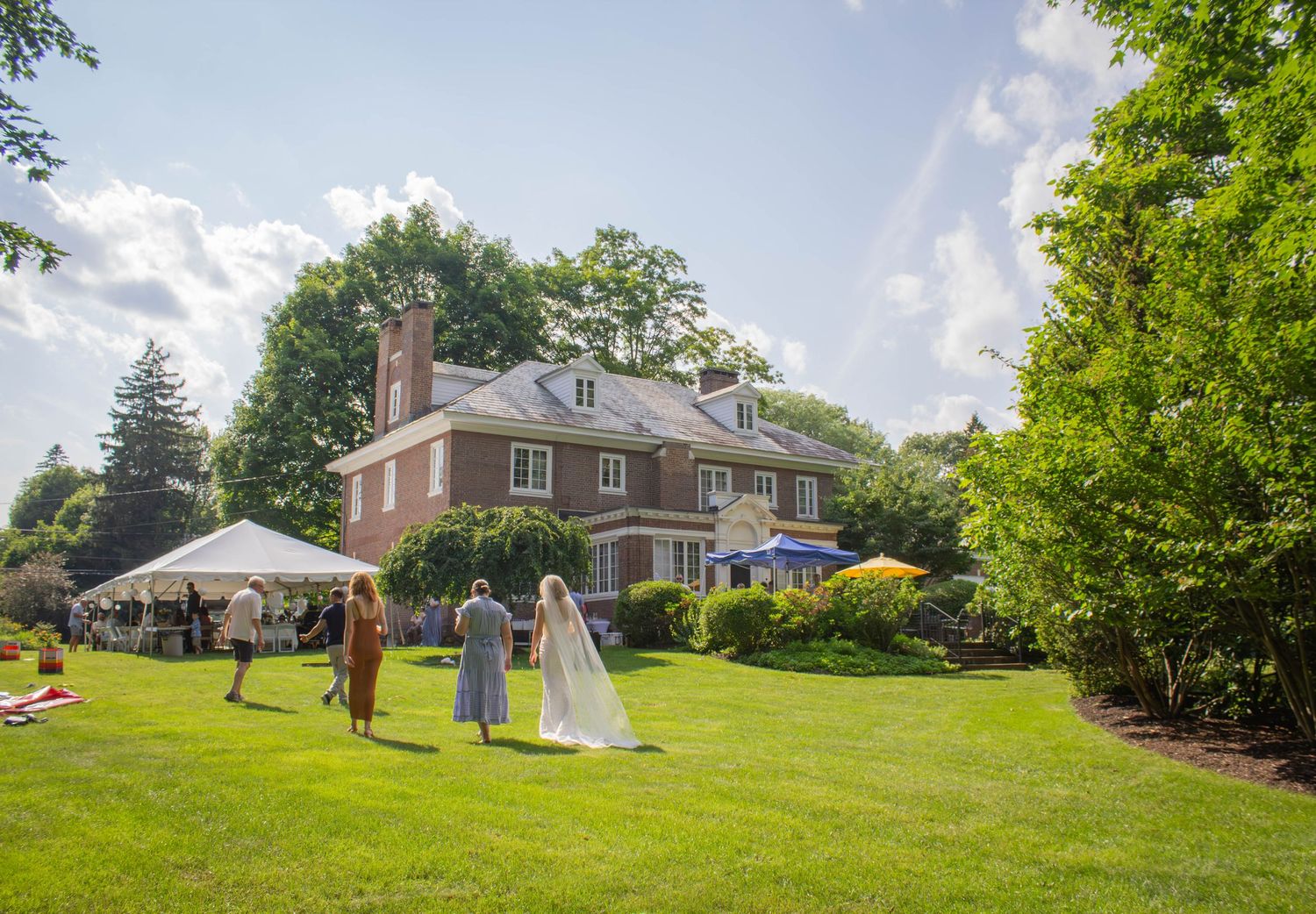 Bride walks across lush green lawn toward elegant brick manor house during outdoor wedding reception.