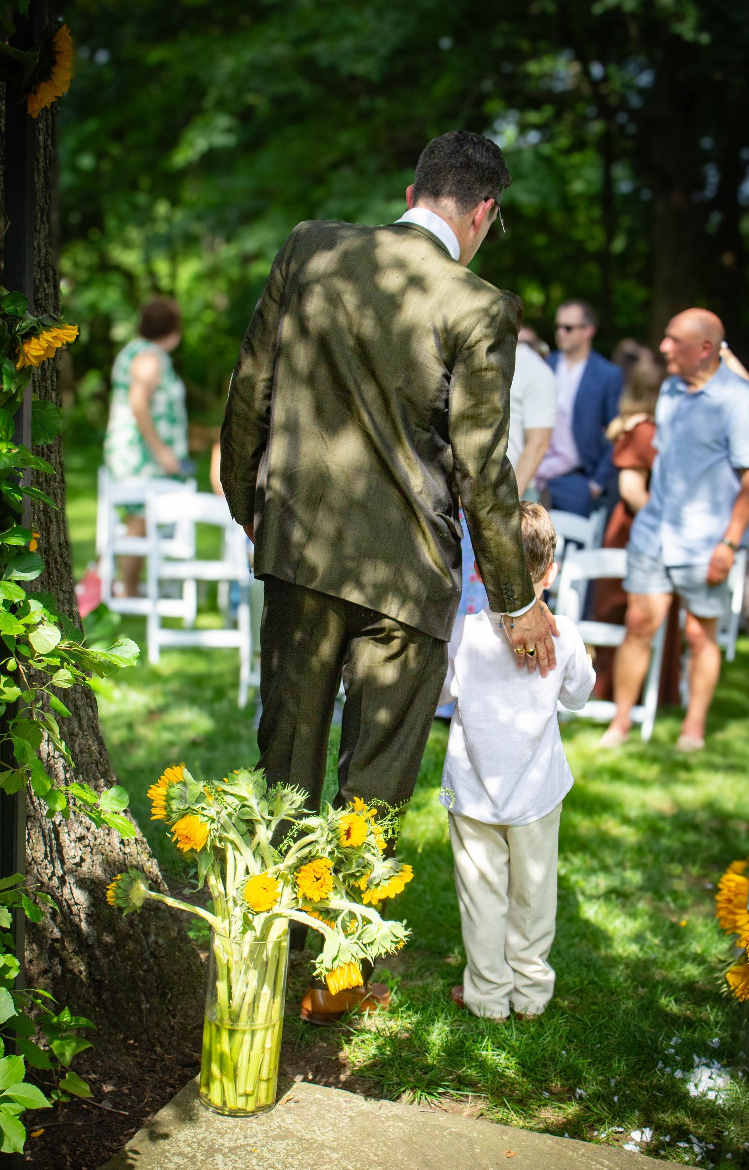 Man in olive suit and child in white stand together at outdoor wedding ceremony with sunflower arrangements.