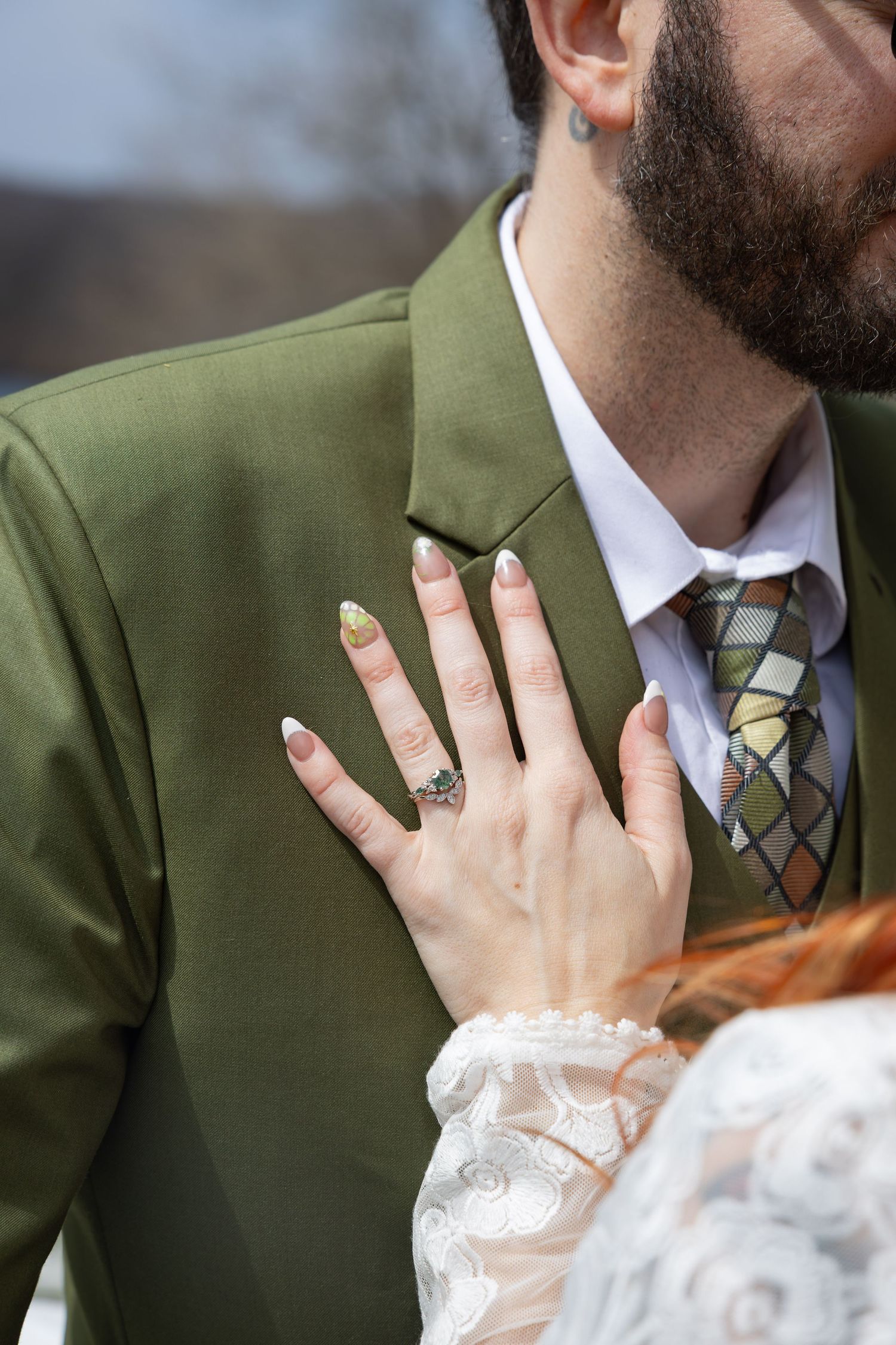 Close-up of bride placing hand on grooms green suit jacket showing engagement ring with styled nails.