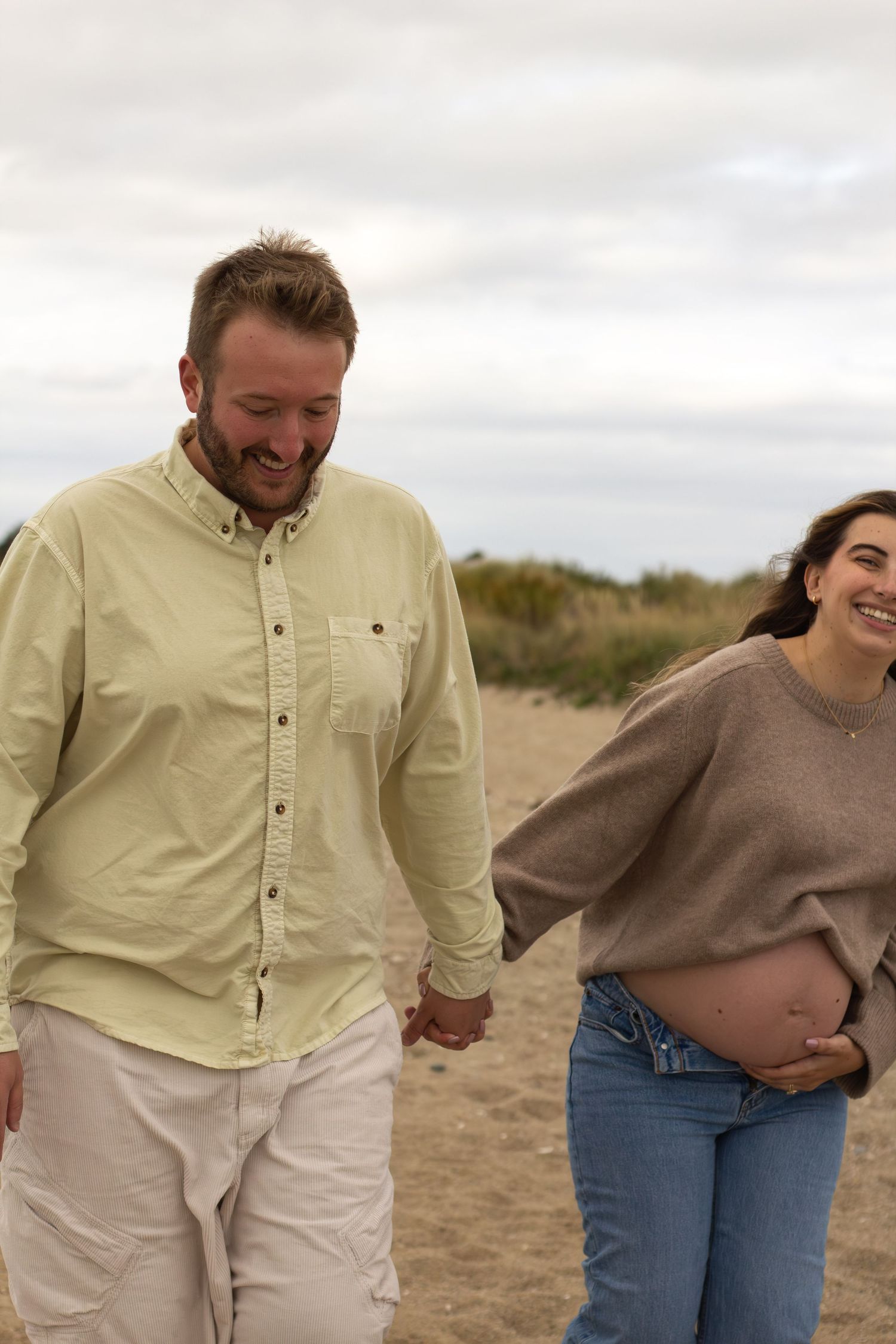 Happy pregnant woman holding baby bump while walking hand-in-hand with partner on a sandy beach path.