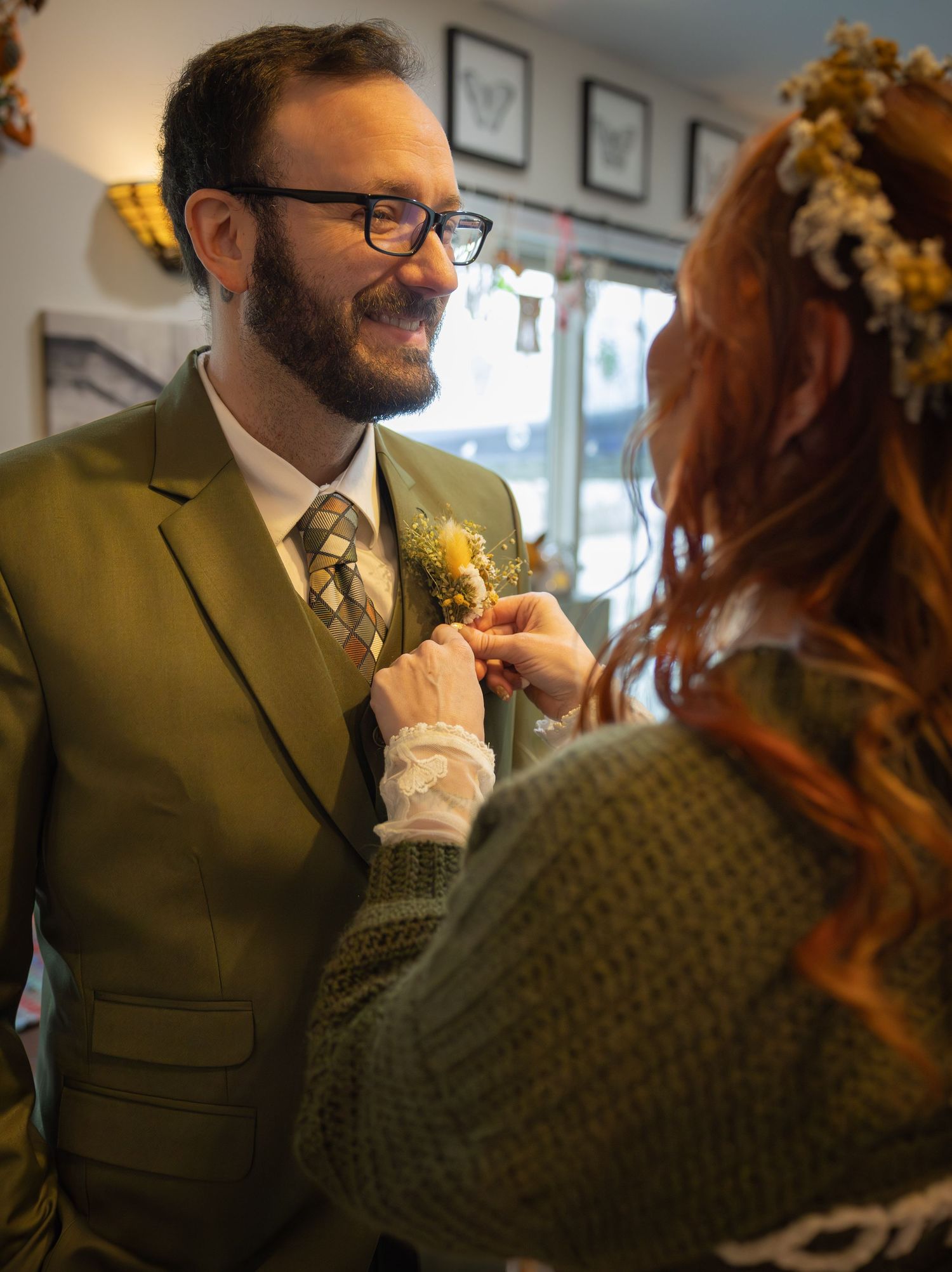 Smiling groom in olive suit having boutonniere pinned by bride wearing floral crown.