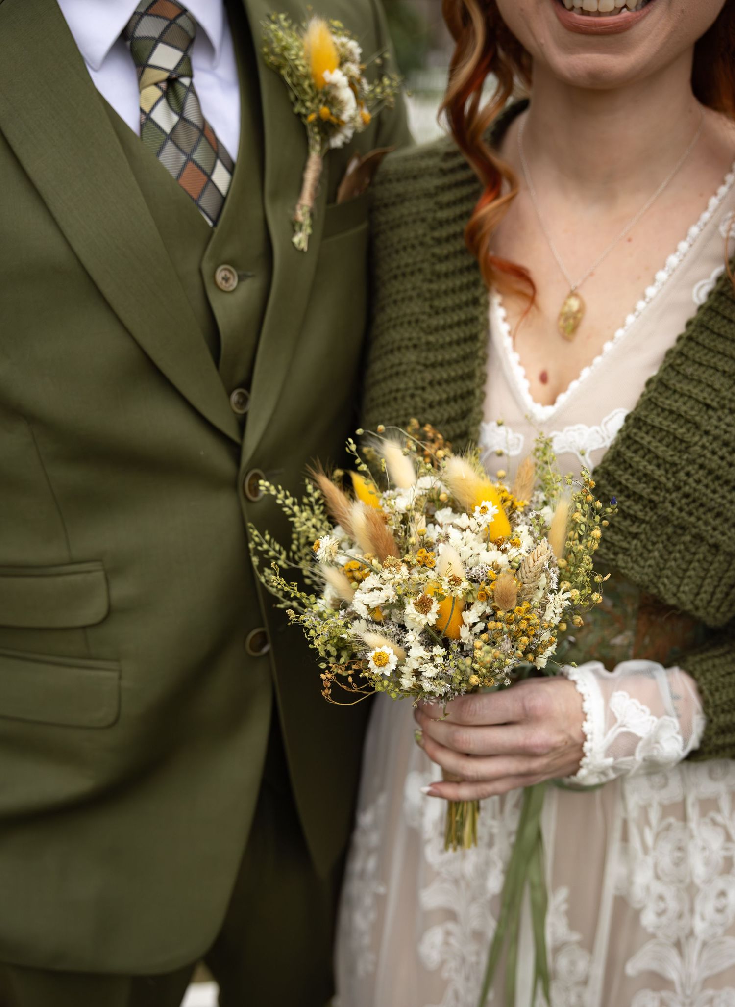 Close-up of bride holding dried wildflower bouquet with yellow blooms beside groom in olive green suit.