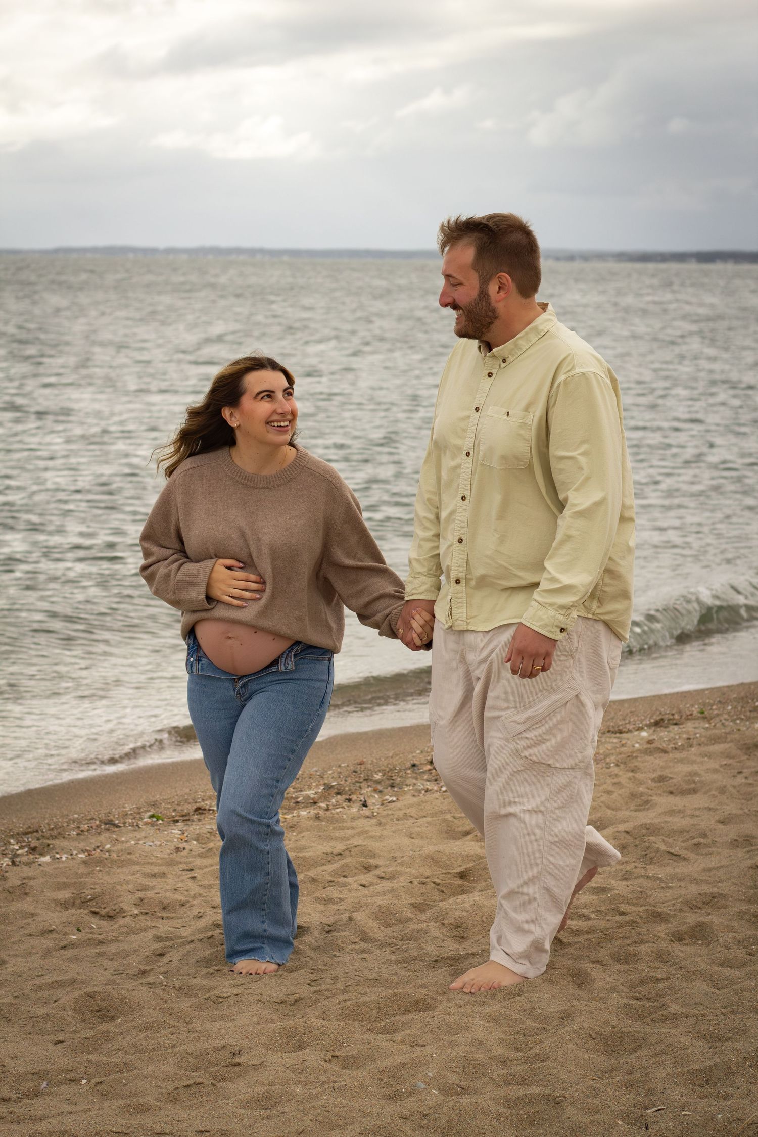 Pregnant couple holding hands and smiling while walking barefoot on a sandy beach by the ocean.